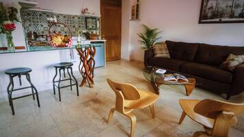 A living room with a brown sofa, two wooden chairs, a glass coffee table, and a kitchen area with a tiled backsplash and a wooden door.