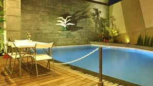 An outdoor swimming pool with a wooden deck, a table and chairs, and potted plants. - Andelir Hotel (Bandung)