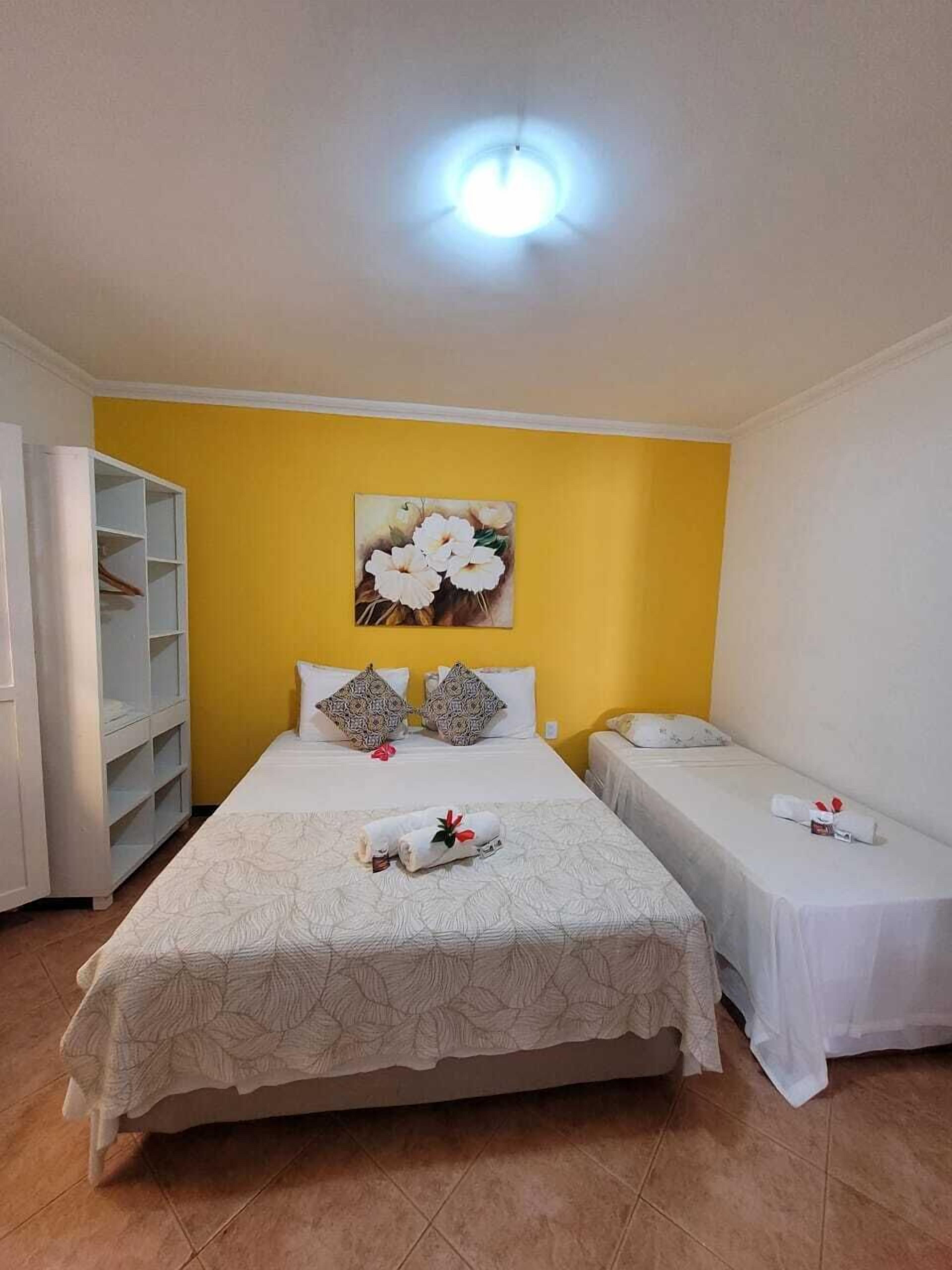 Quarto de hotel com uma cama grande, duas mesinhas de cabeceira, um guarda-roupa e um quadro com flores na parede.