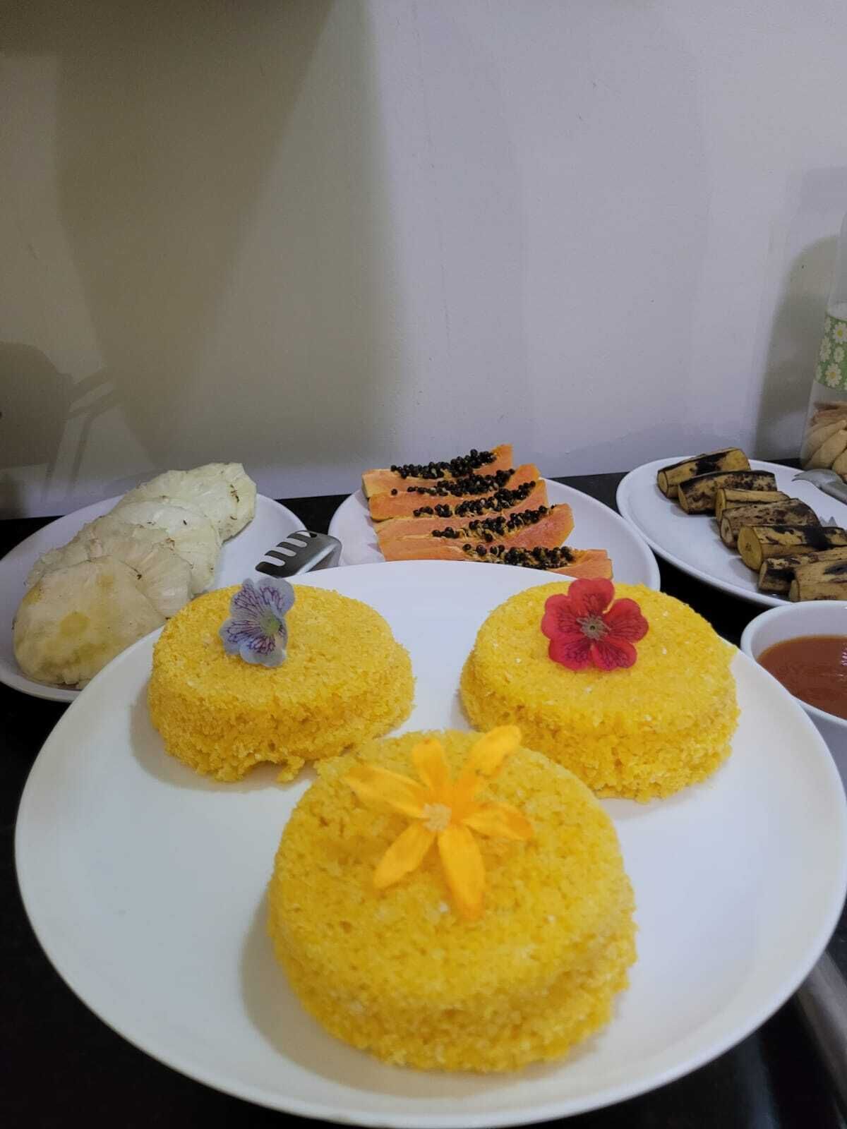 Uma mesa com diversos pratos, incluindo arroz amarelo decorado com flores, um prato branco com uma flor roxa e um prato com fatias de laranja e sementes pretas.