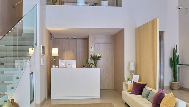 A modern reception area with a white reception desk, a sofa with colorful cushions, a large wooden planter, and a glass staircase. - Hotel Omnium (Barcelona)