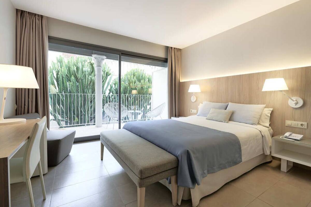 Habitación de hotel con cama, mesitas de noche, escritorio con silla, balcón y vistas a cactus, además de un ventanal grande.