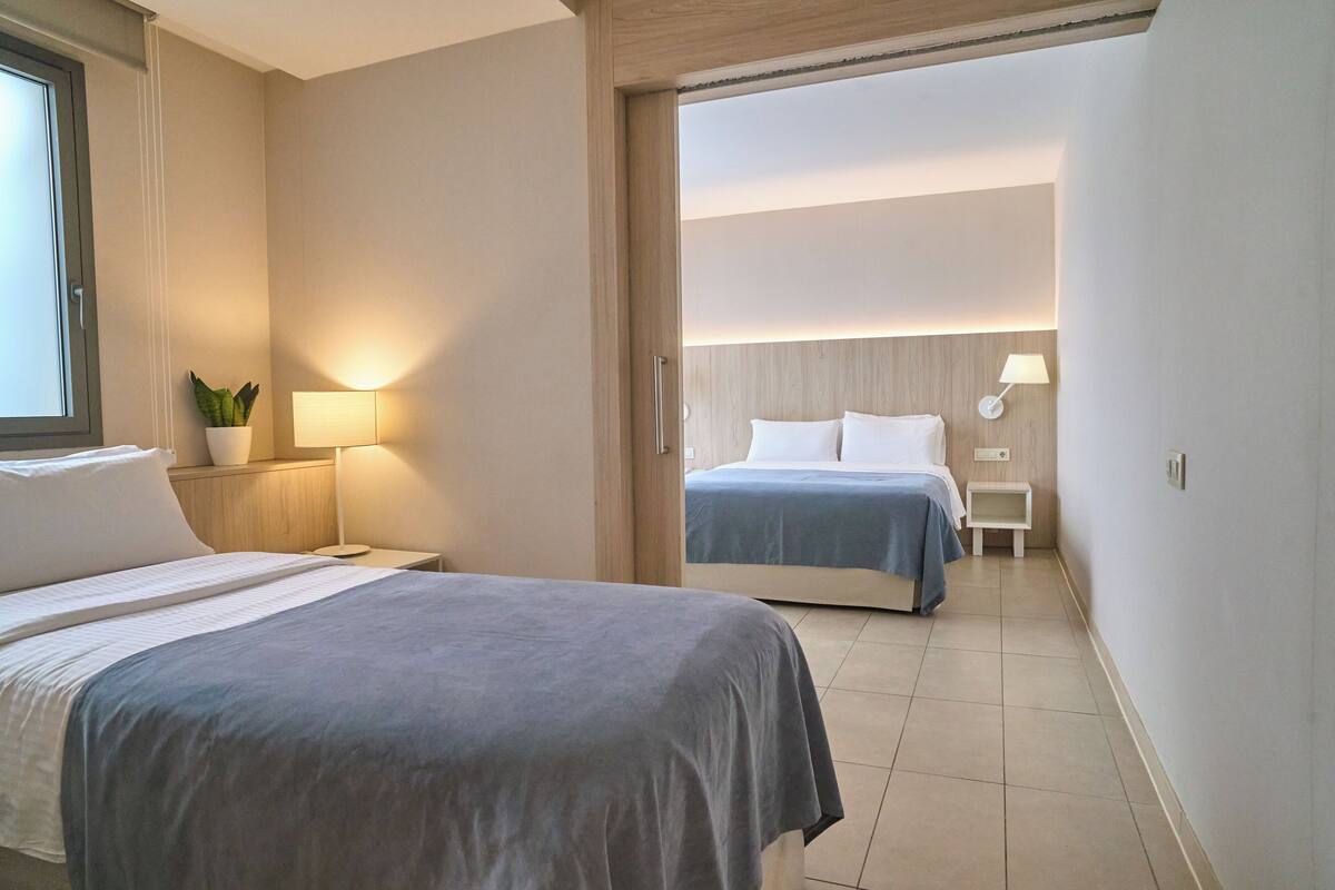 Habitación de hotel con dos camas, una mesita de noche con lámpara, una planta pequeña y vista a otra habitación.