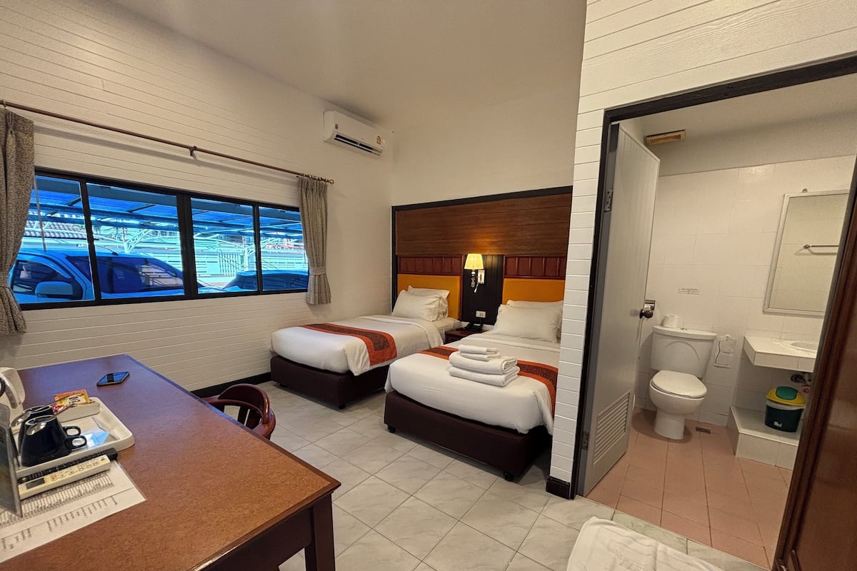 A hotel room with two beds, a desk with a chair, a window with a view of the outside, and a bathroom with a toilet and sink.