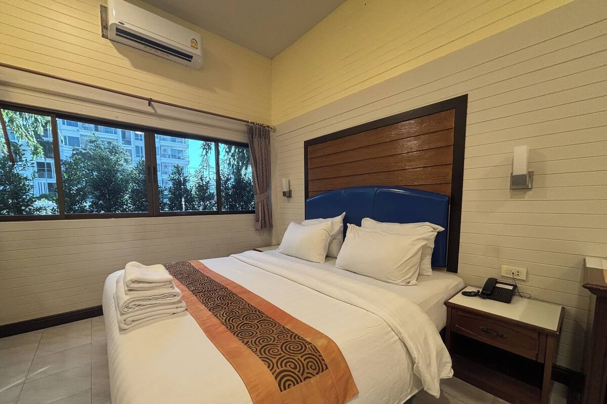 A hotel room with a bed, a bedside table with a phone, a window with curtains, and an air conditioning unit.