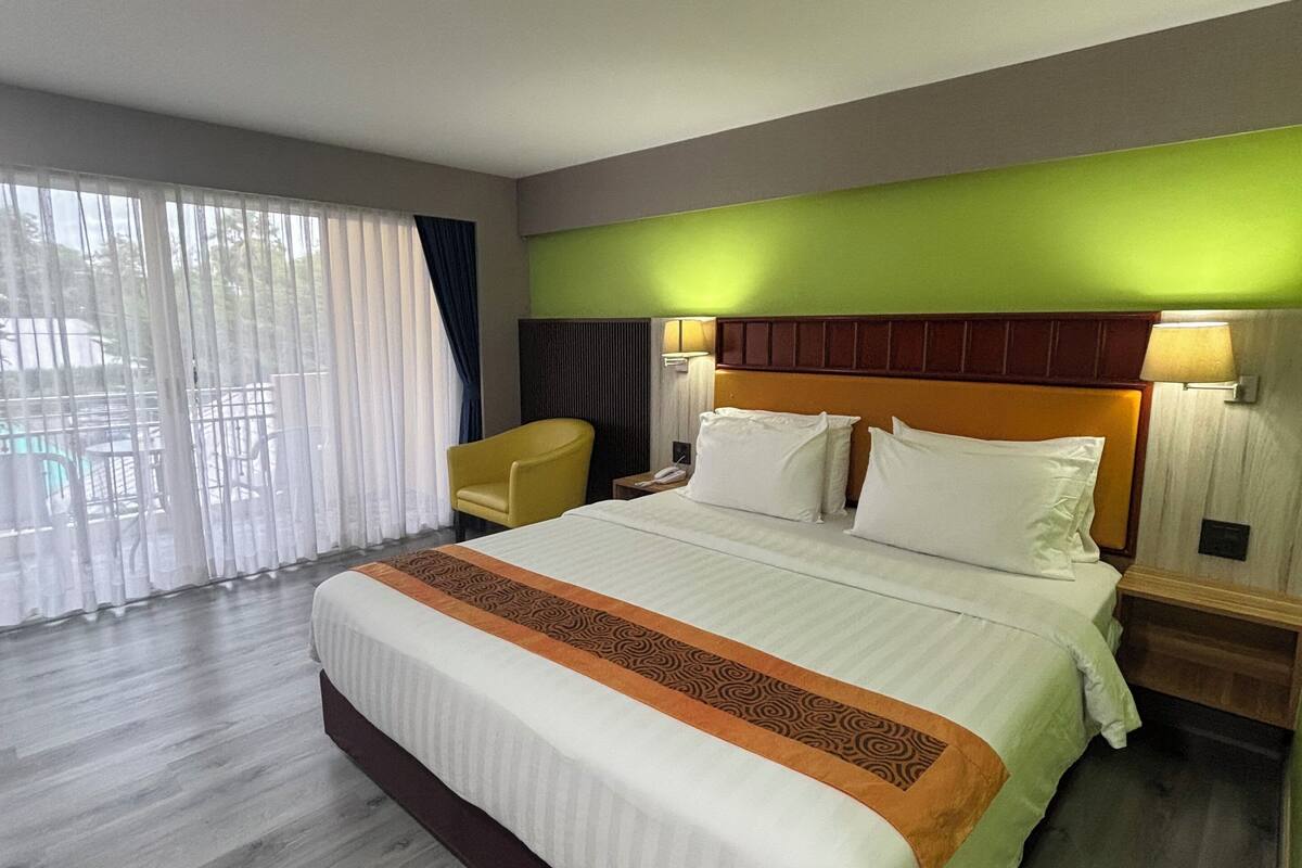 A hotel room with a large bed, a wooden floor, a green accent wall, and a view through a large window.