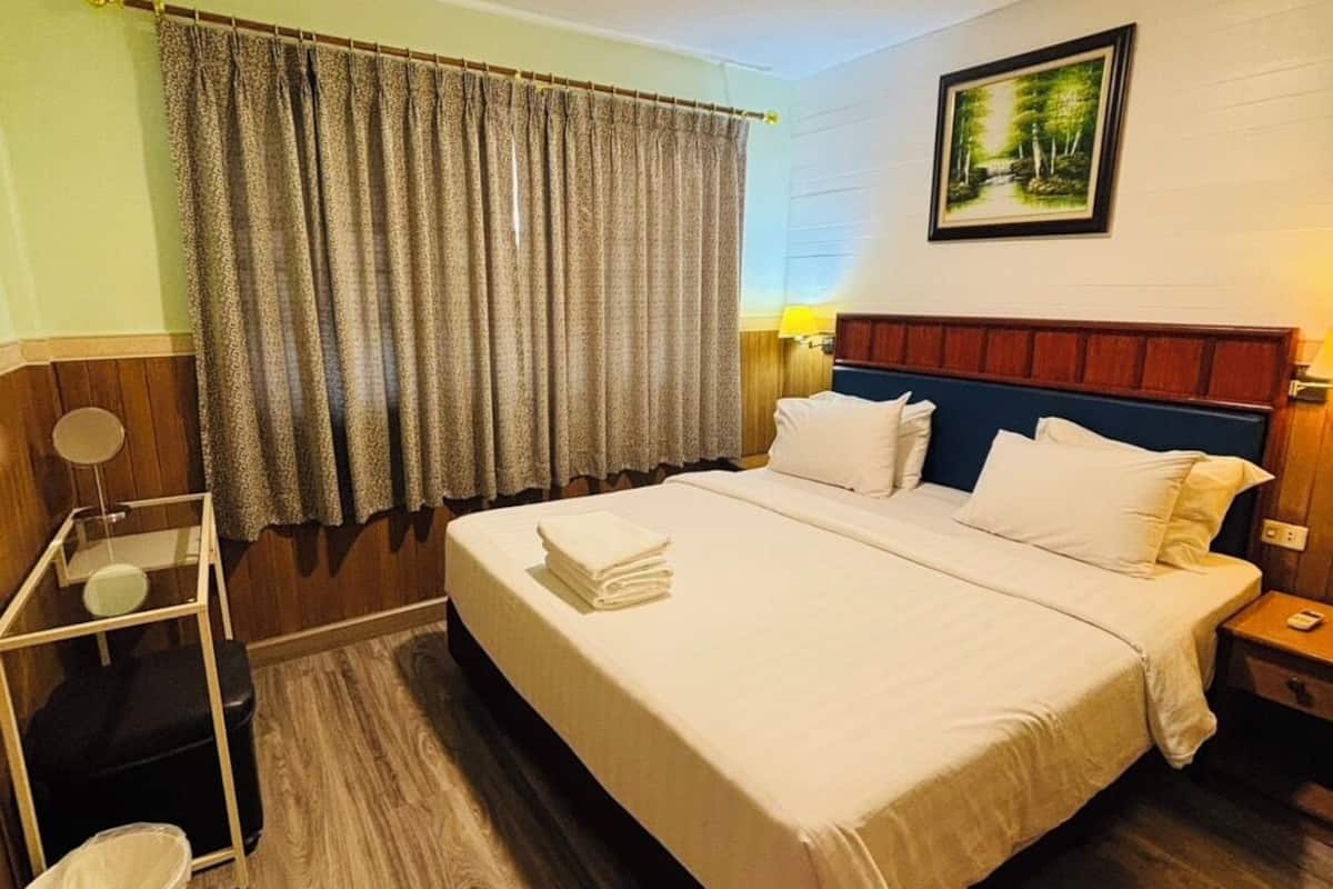 A hotel room with a large bed, a wooden floor, a curtain, a nightstand, a lamp, and a painting on the wall.