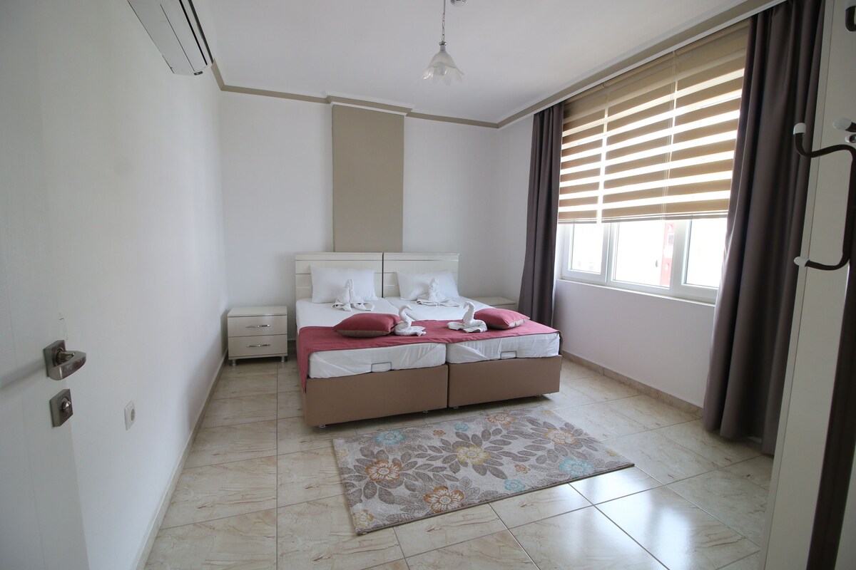 A bedroom with a bed, a bedside table, a nightstand, a window with blinds, and a door.