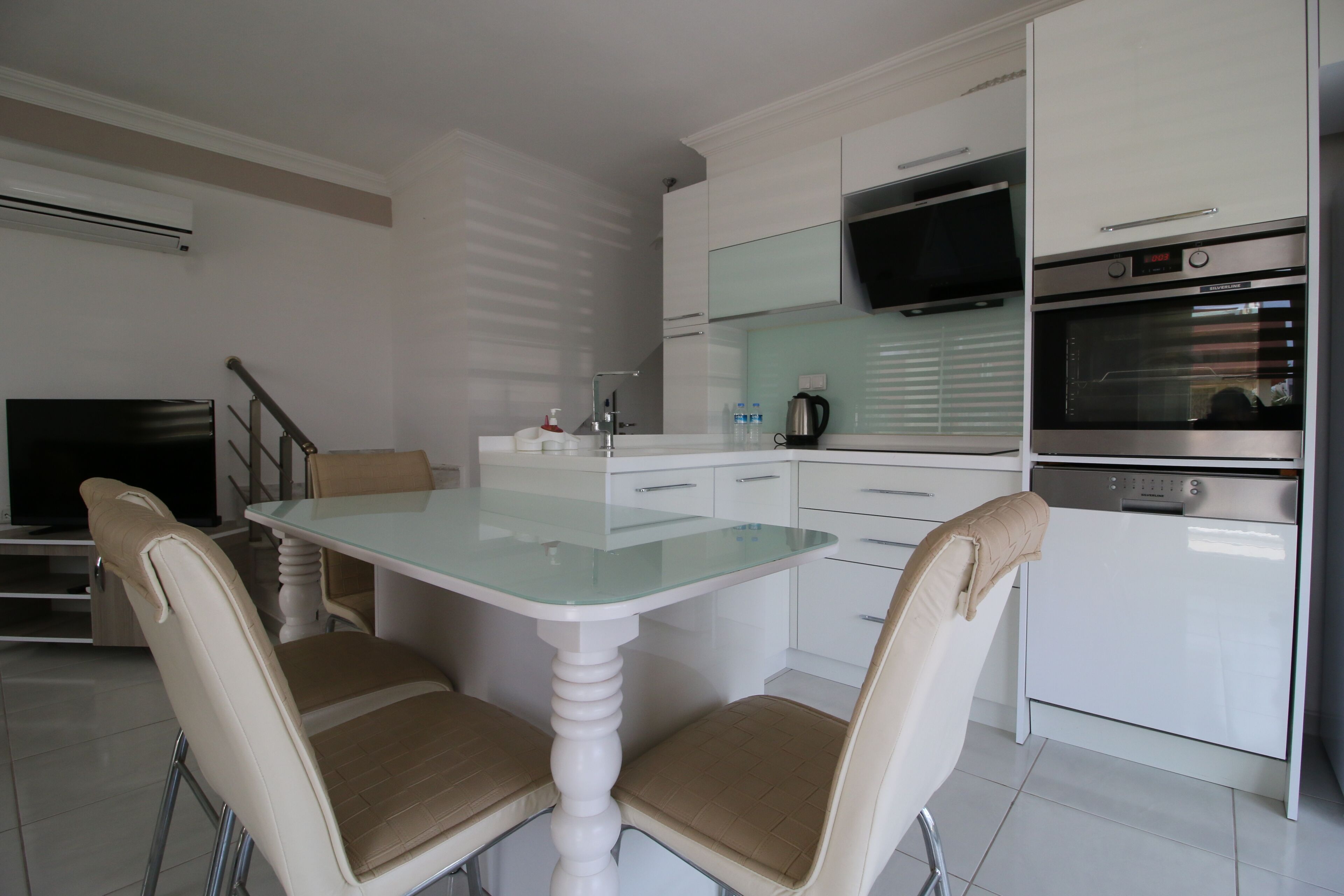 A modern kitchen with white cabinetry, a glass-top dining table, two beige chairs, a built-in oven, and a flat-screen TV.
