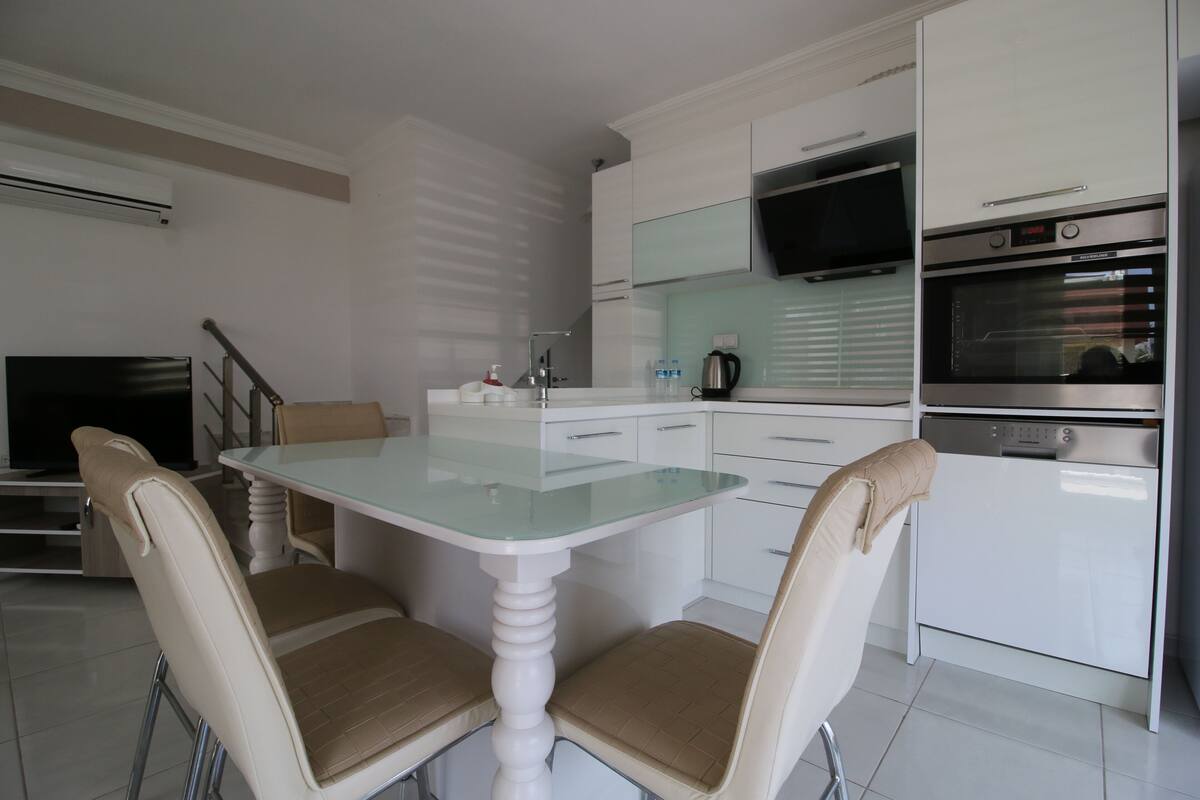 Een moderne keuken met witte kasten, een eettafel met glazen blad, twee beige stoelen, een inbouwoven en een flatscreen televisie.