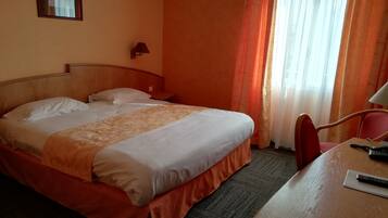 A hotel room with a bed, a desk, a chair, a lamp, and a window with curtains.