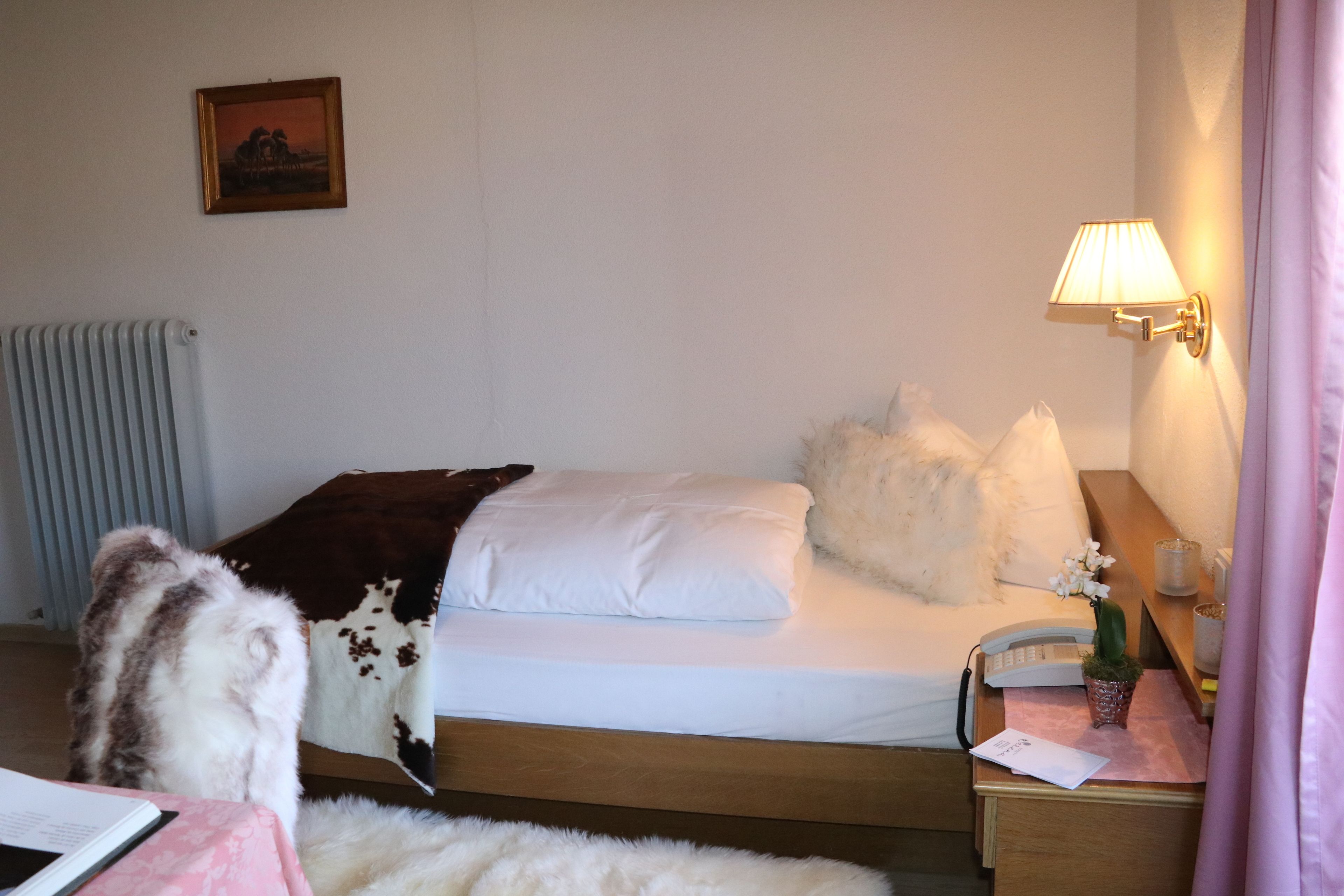Une chambre d’hôtel avec un lit, une lampe de chevet, un téléphone, une petite table sur laquelle est posé un vase de fleurs et un radiateur.