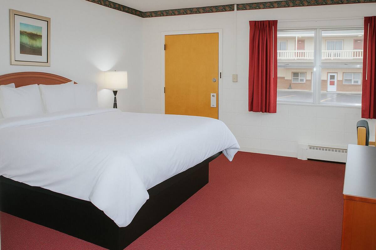 A hotel room with a large bed, a wooden headboard, a bedside lamp, a red carpet, a window with red curtains, and a view of a building outside.