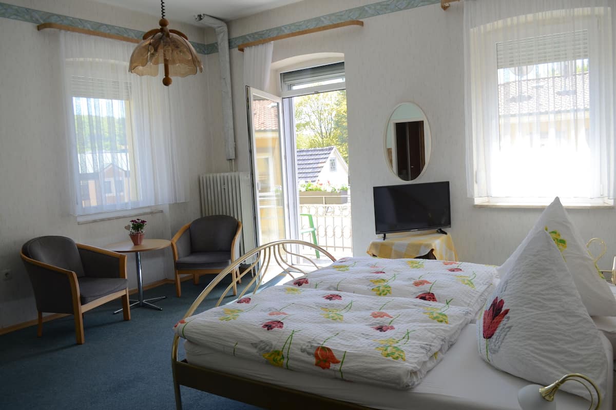 Quarto com cama, duas cadeiras, mesa pequena com planta, televisão e janela com cortinas.