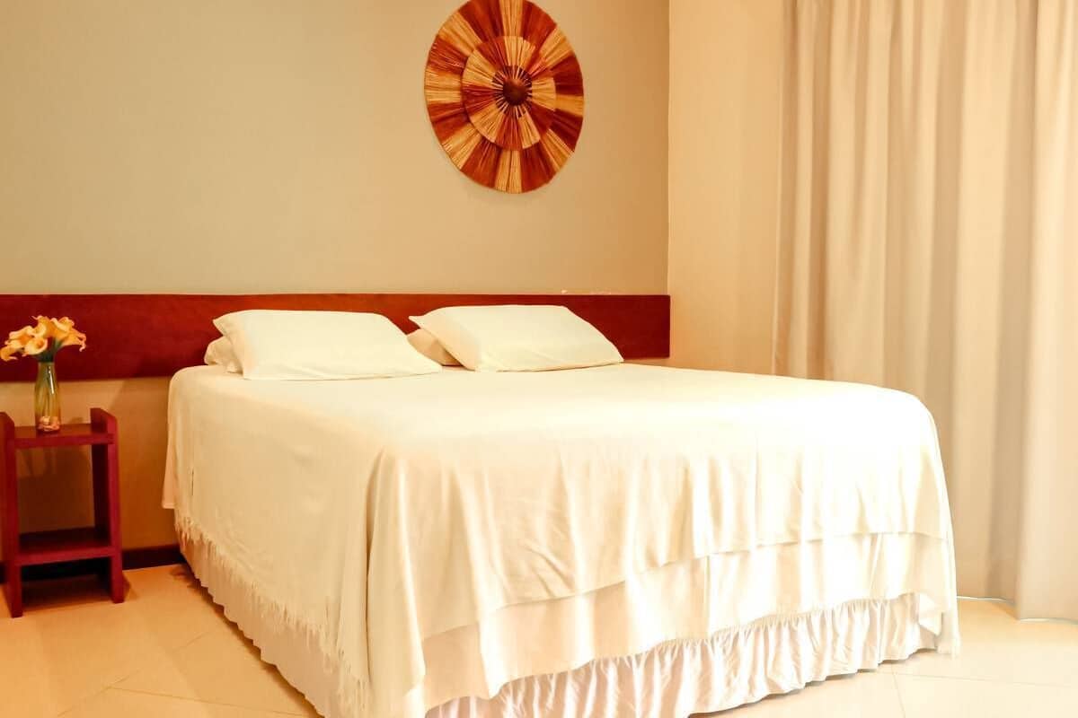 Una cama bien hecha con ropa blanca, una mesita de noche de madera con un jarrón de flores y una obra de arte en la pared sobre la cama.