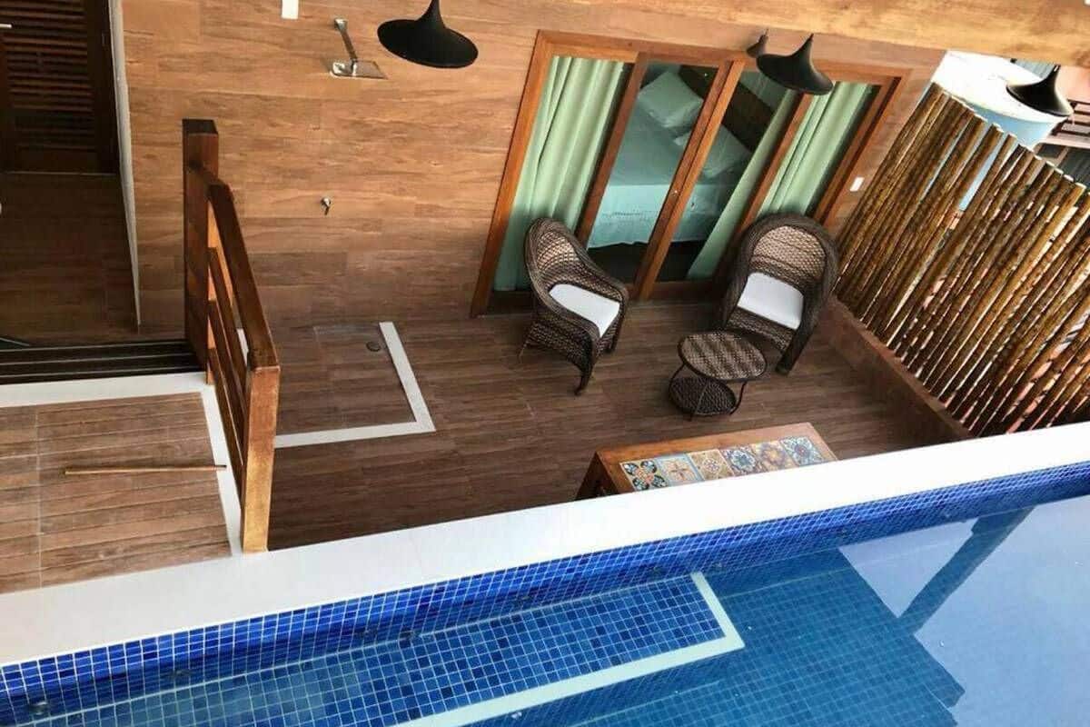 Un área de piscina interior moderna con suelo de madera, dos sillas de mimbre, una pequeña mesa y un vidrio que da a una habitación.