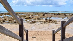 Un escalier en bois mène à une plage de sable, d’où l’on peut admirer la mer et apercevoir un bateau au loin.