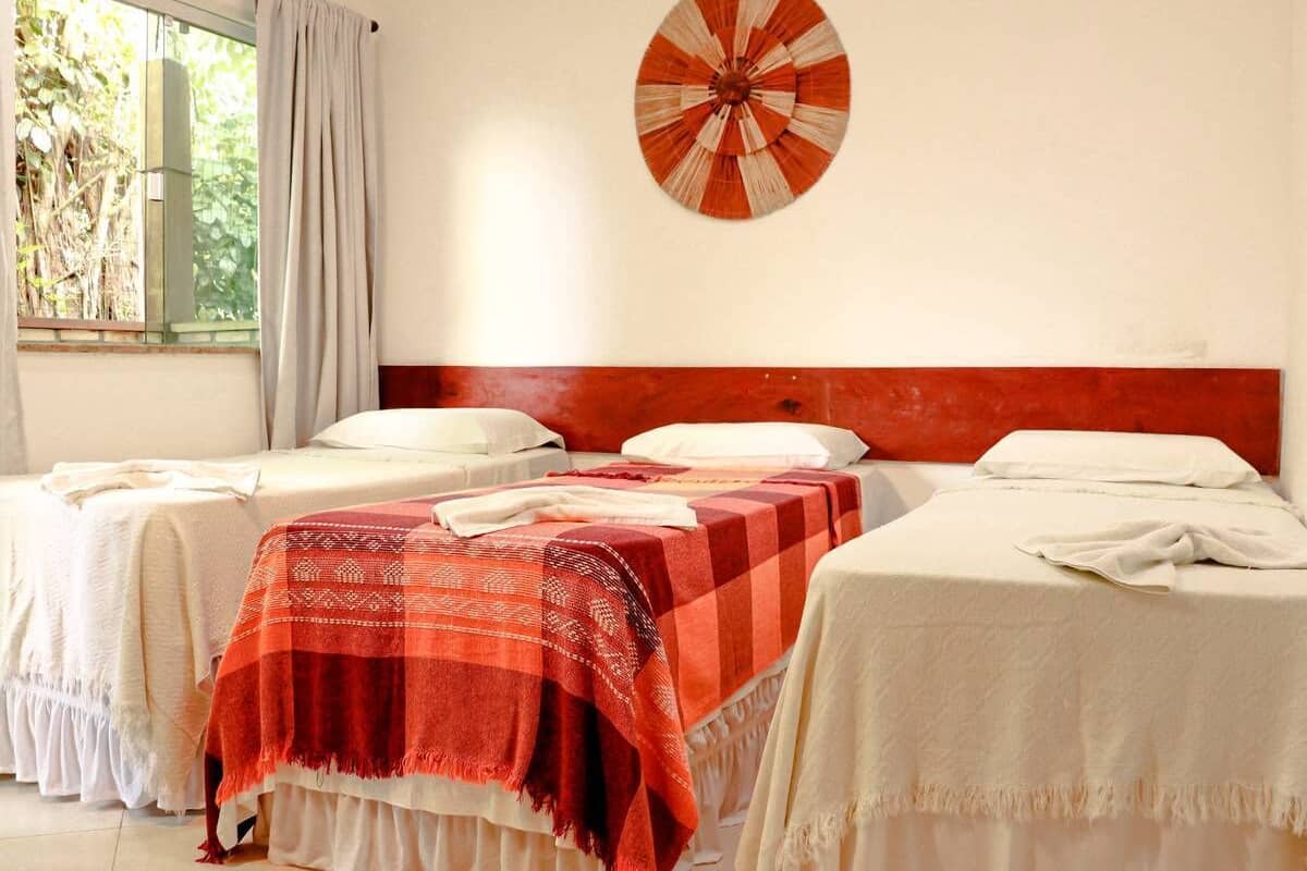 Una habitación de hotel con dos camas individuales, una con una colcha roja y blanca a cuadros y la otra con un edredón blanco liso. Hay un dardo en la pared y una ventana con cortinas.