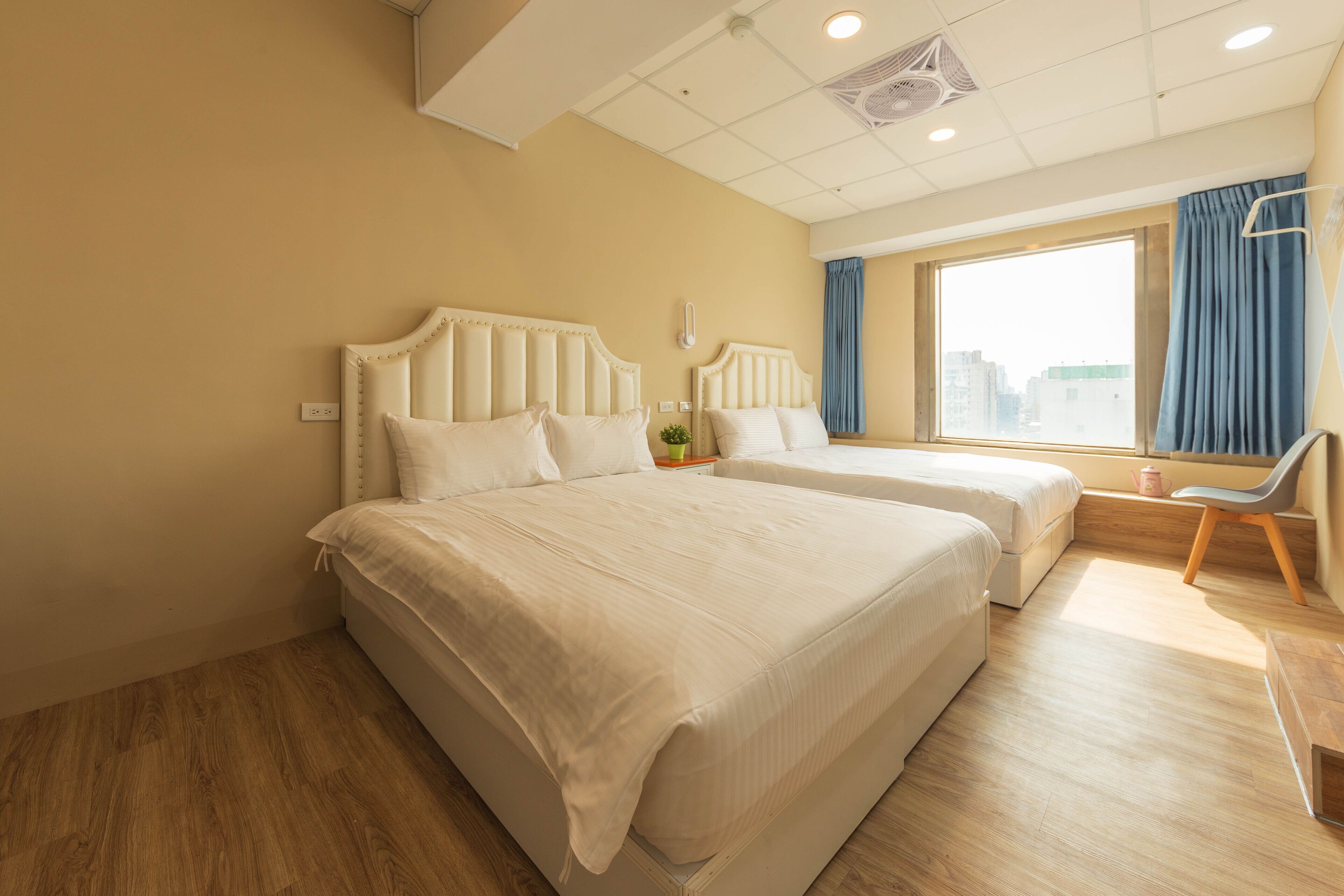 A hotel room with two beds, a large window with curtains, a chair, and a small table.