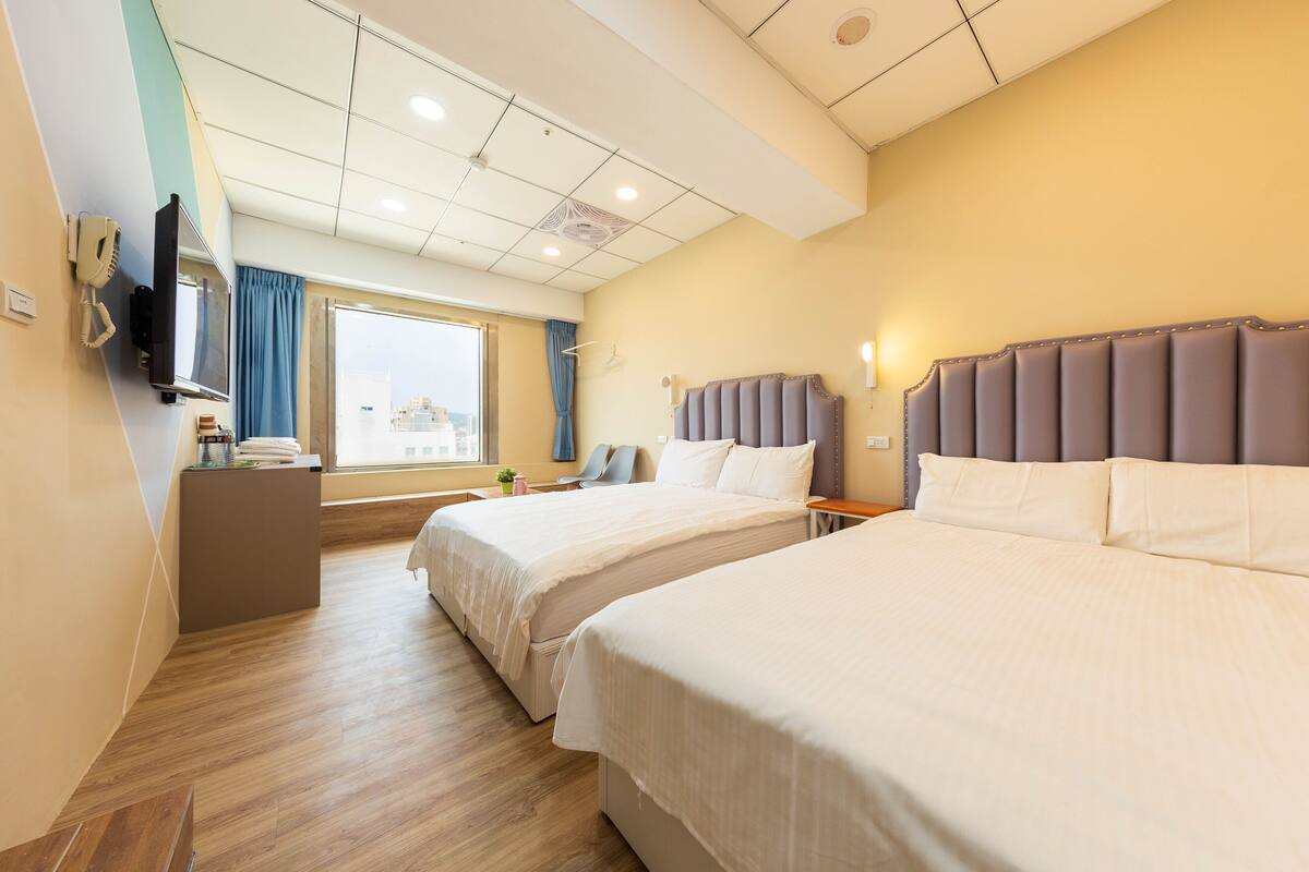 A hotel room with two beds, a desk, a TV, and a window with curtains.