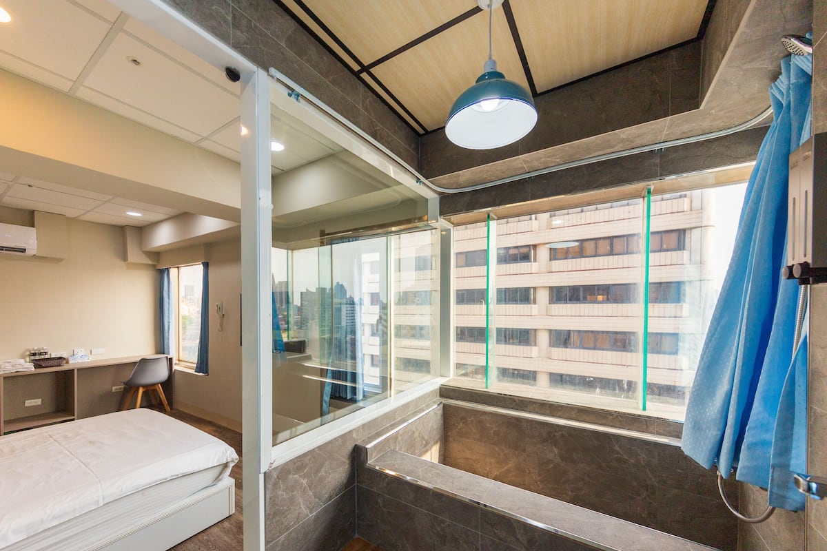A modern hotel room with a bed, a desk, and a large window offering a city view.