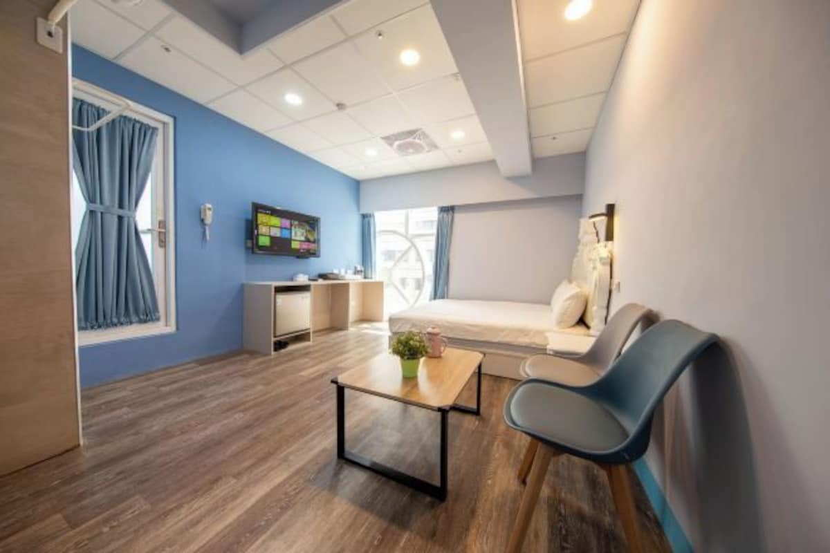 Una habitación de hotel moderna con una pared azul de acento, suelo de madera, cama con cabecera blanca, zona de estar con una silla azul, una mesita con una planta y un televisor en la pared.