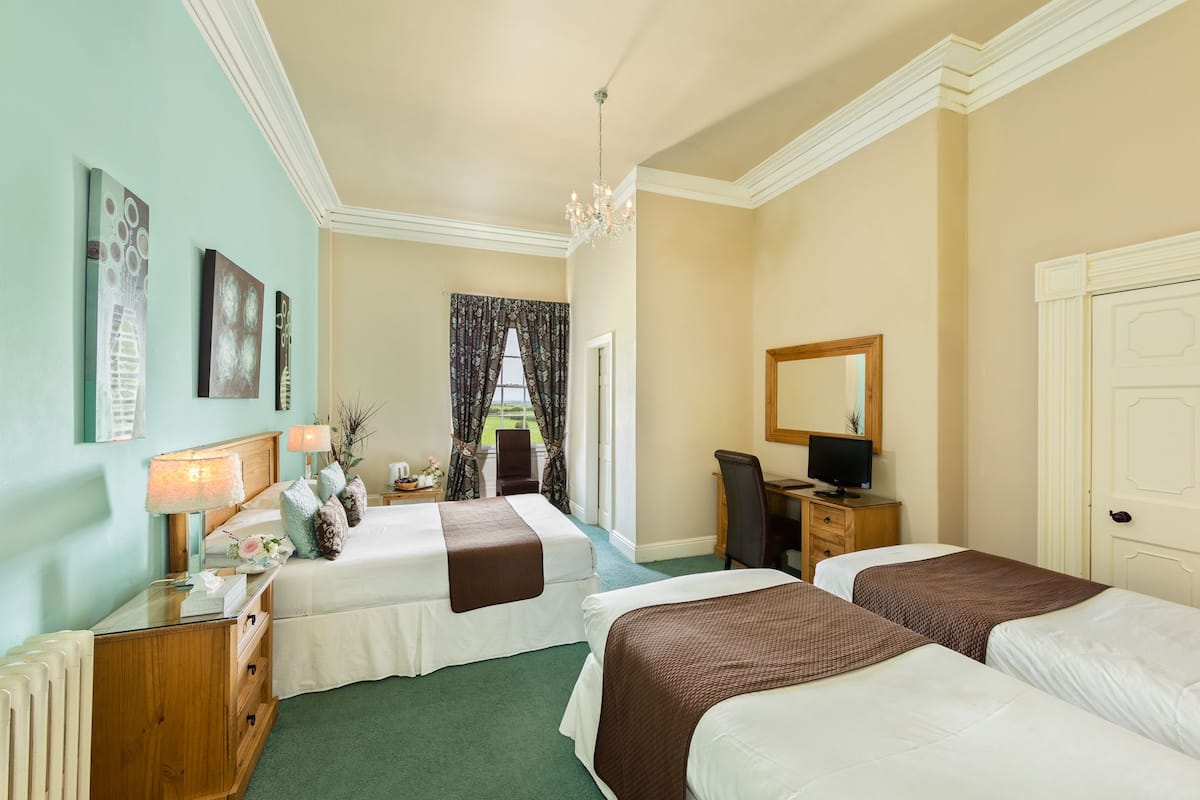 Une chambre d’hôtel avec deux lits, un bureau avec un ordinateur, une chaise, une fenêtre avec des rideaux et des œuvres d’art accrochées aux murs.