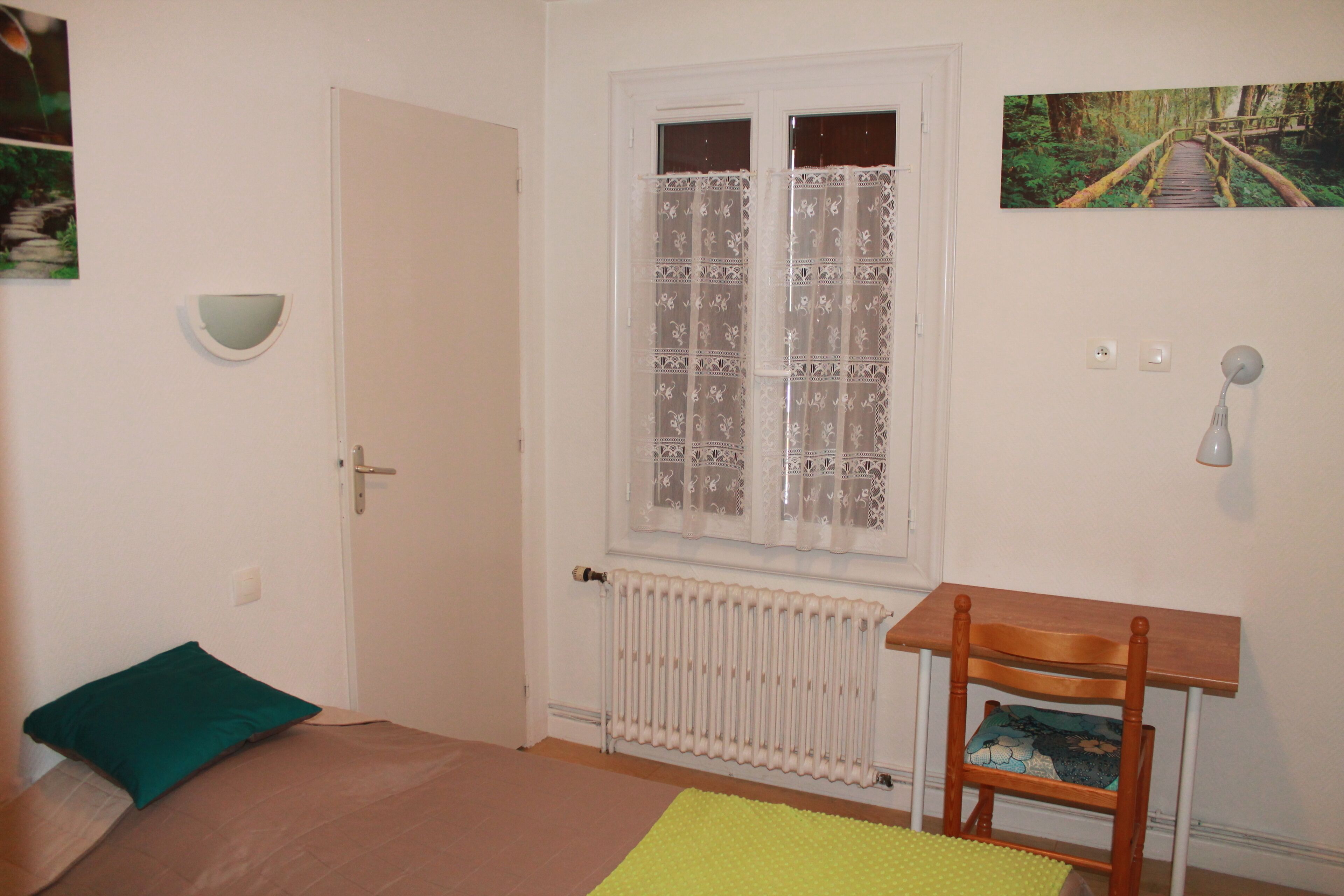 Une pièce comprenant un lit, une chaise, un radiateur, une fenêtre avec des rideaux et une lampe fixée au mur.