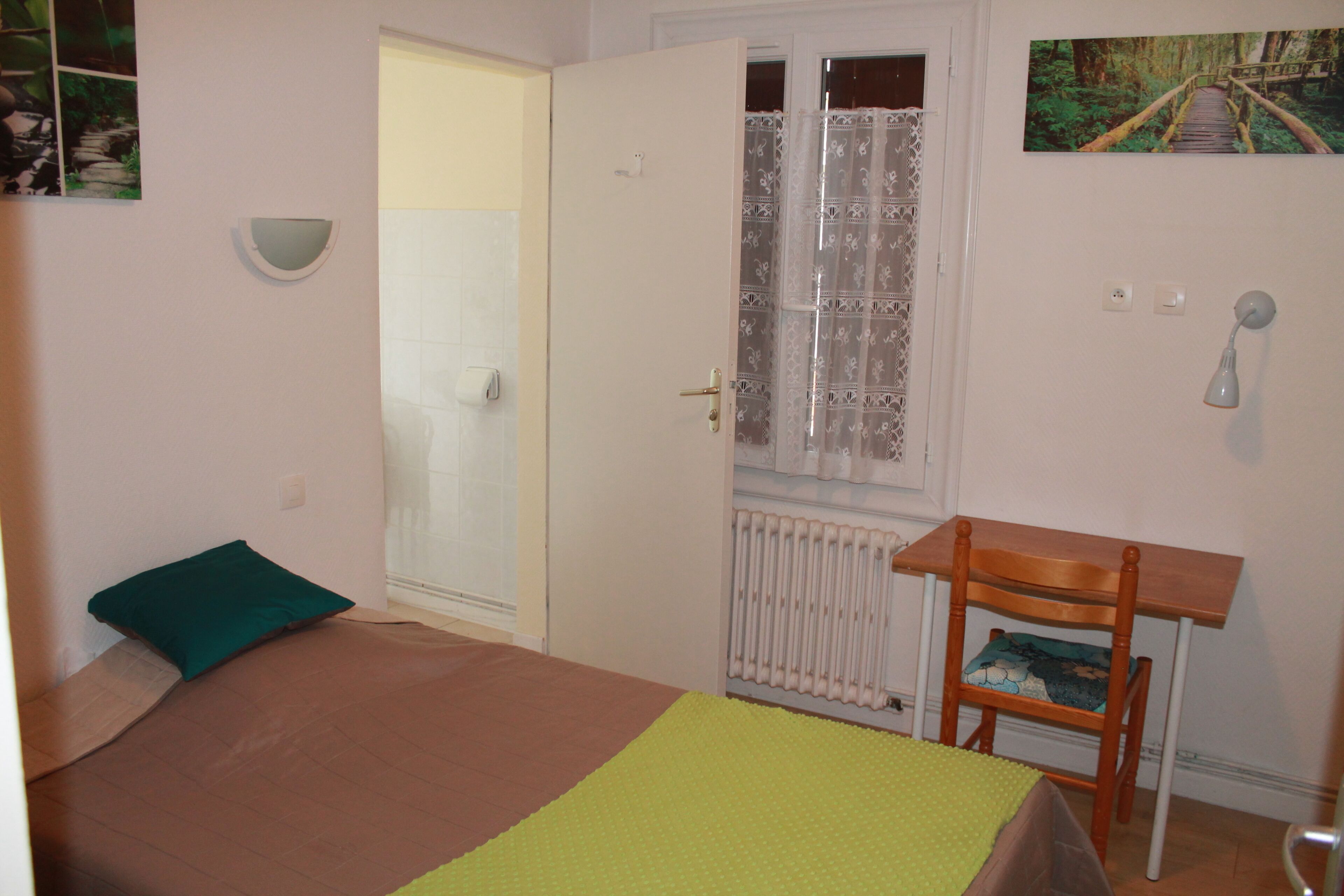 Quarto com cama, janela com cortina, radiador, cadeira de madeira e uma pequena mesa.