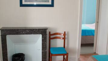 Une chambre avec une chaise en bois, un tableau encadré au mur, un lit recouvert d’une couverture bleue et une porte donnant sur une autre pièce.