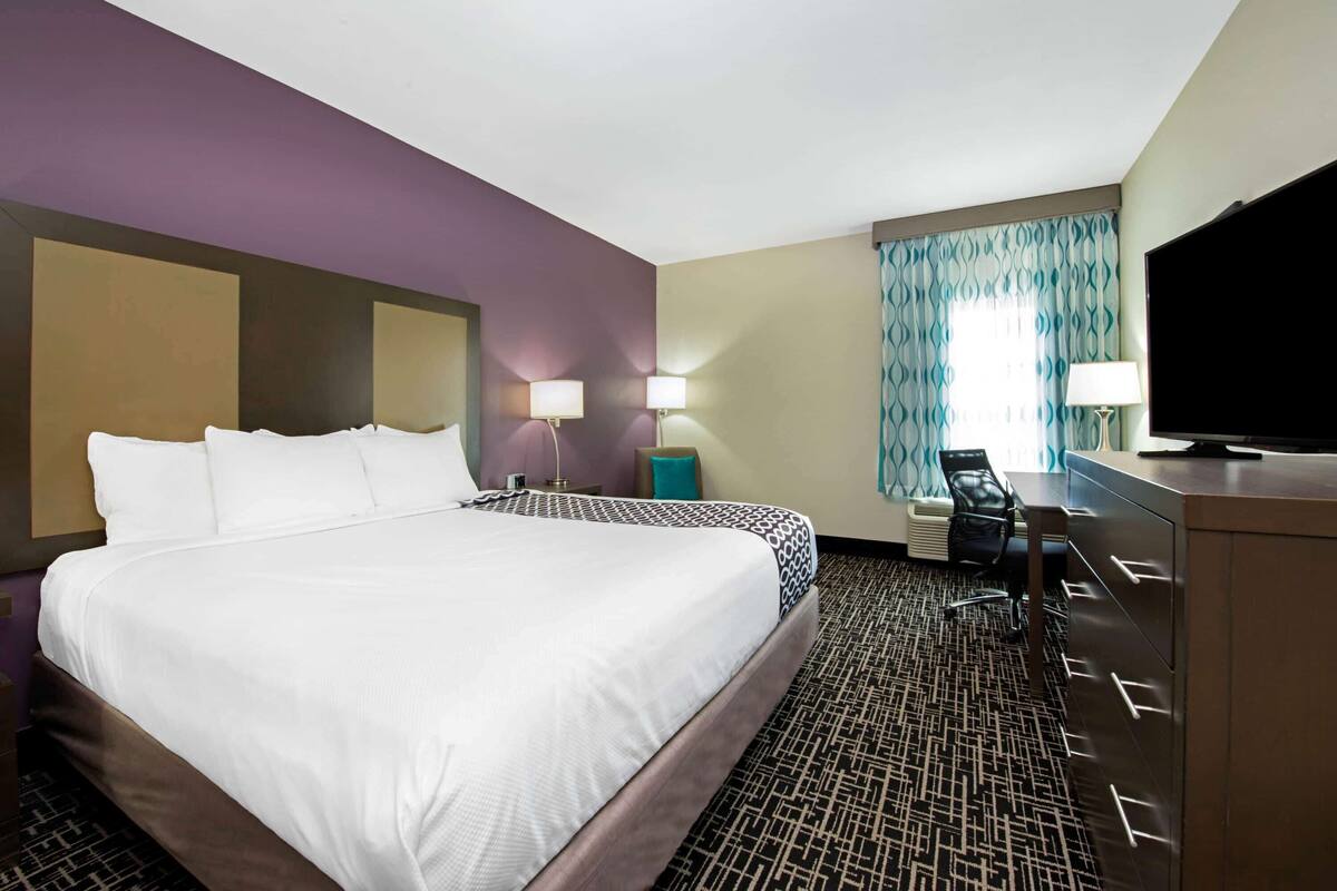 A hotel room with a large bed, a desk with a flat-screen TV, and a window with curtains.