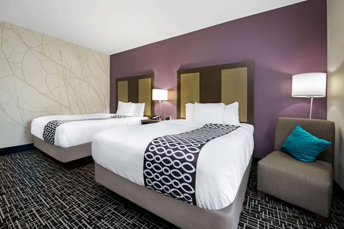 A hotel room with two beds, a small armchair, and a nightstand with a lamp.