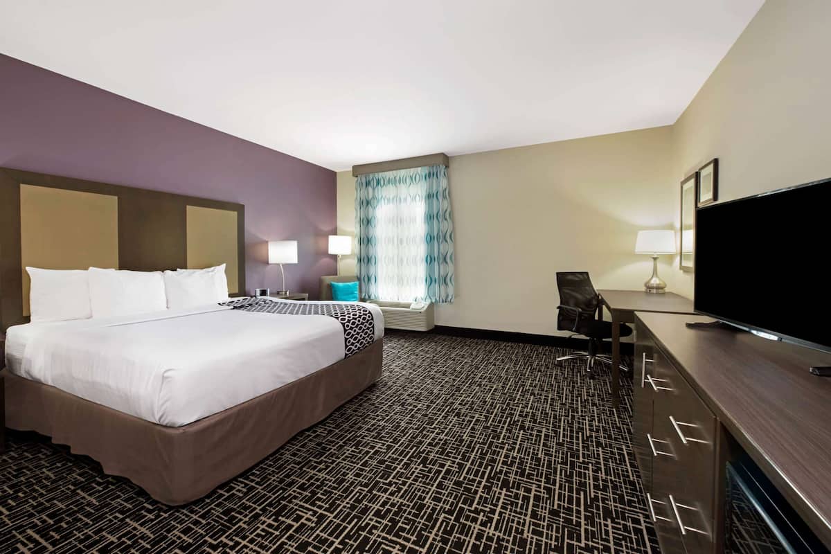 A hotel room with a large bed, a desk with a flat-screen TV, a chair, a desk lamp, and a window with curtains.