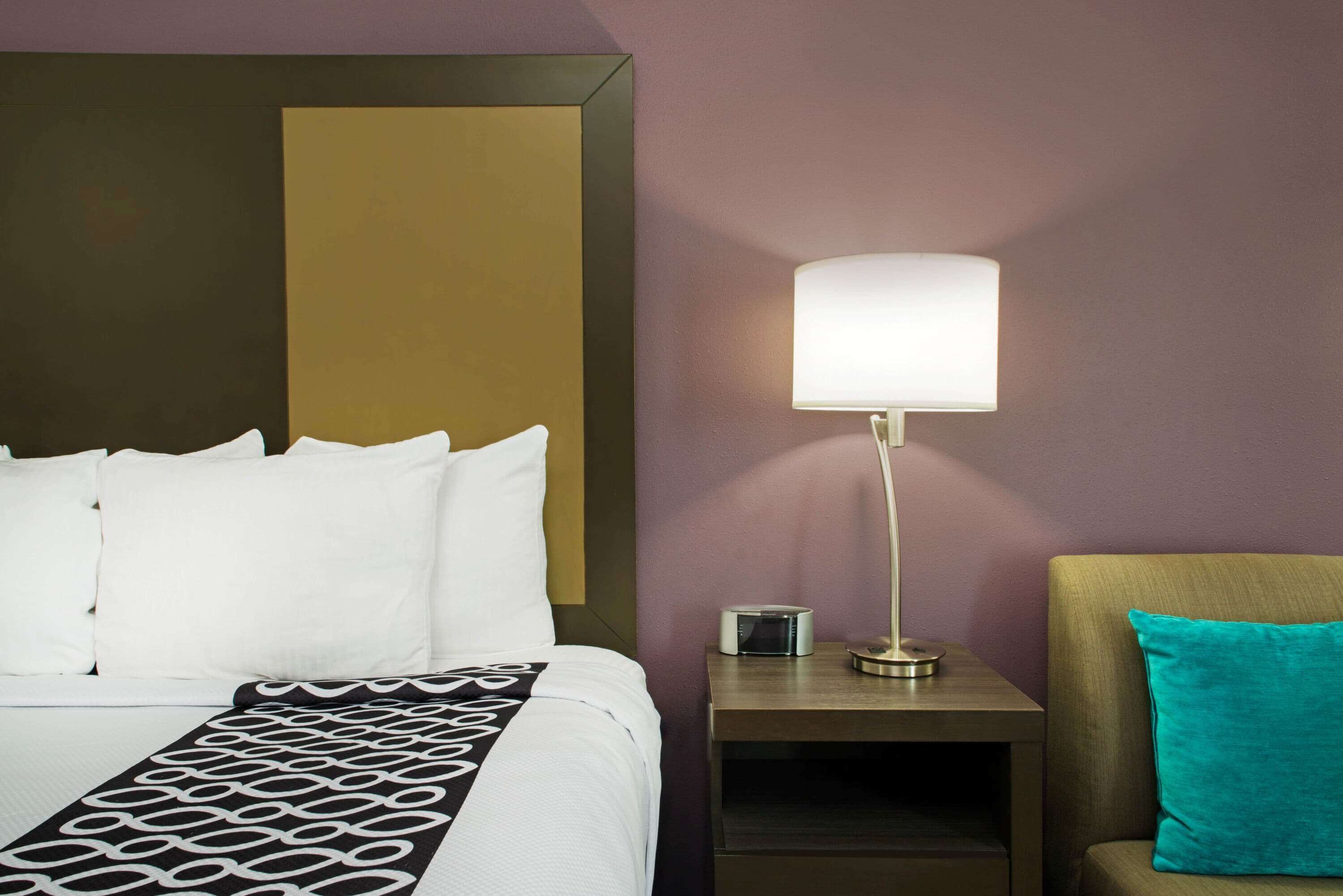 A hotel room with a bed, a bedside table with a lamp, and a chair with a teal cushion.
