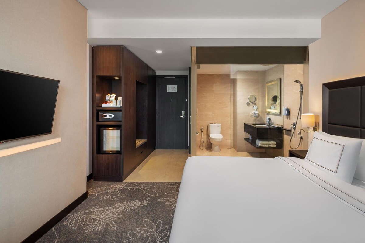 A hotel room with a bed, a TV, a minibar, a bathroom with a toilet and sink, and a shower.