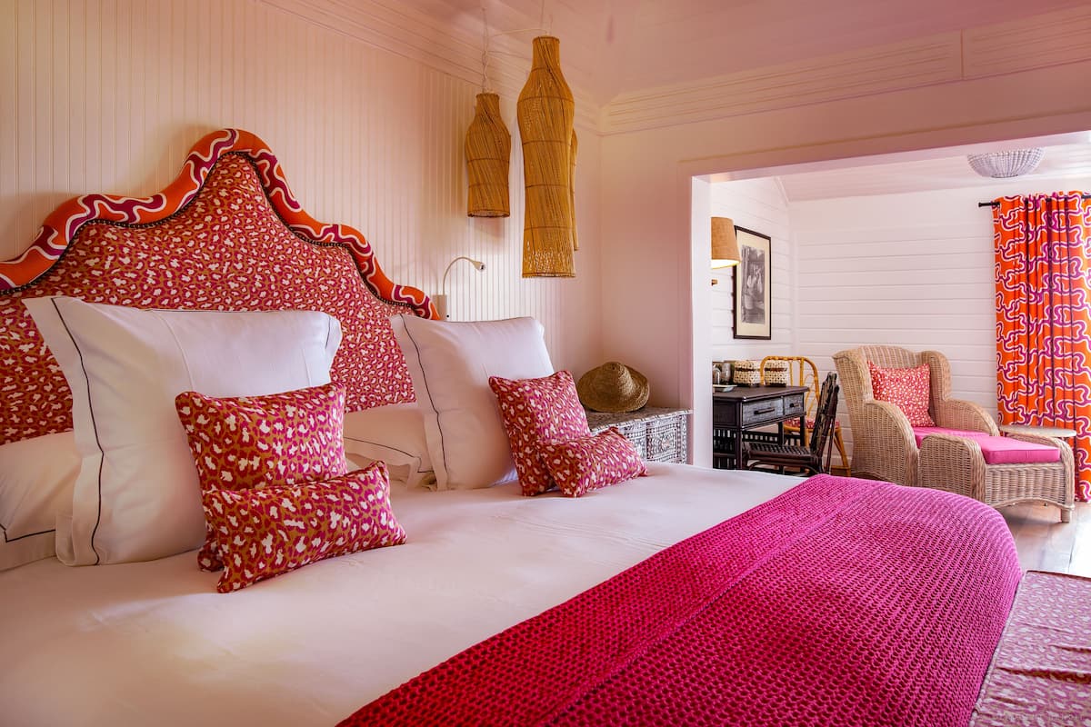 Un dormitorio con una cama grande, varias almohadas, una manta roja, muebles de mimbre y vistas al exterior.
