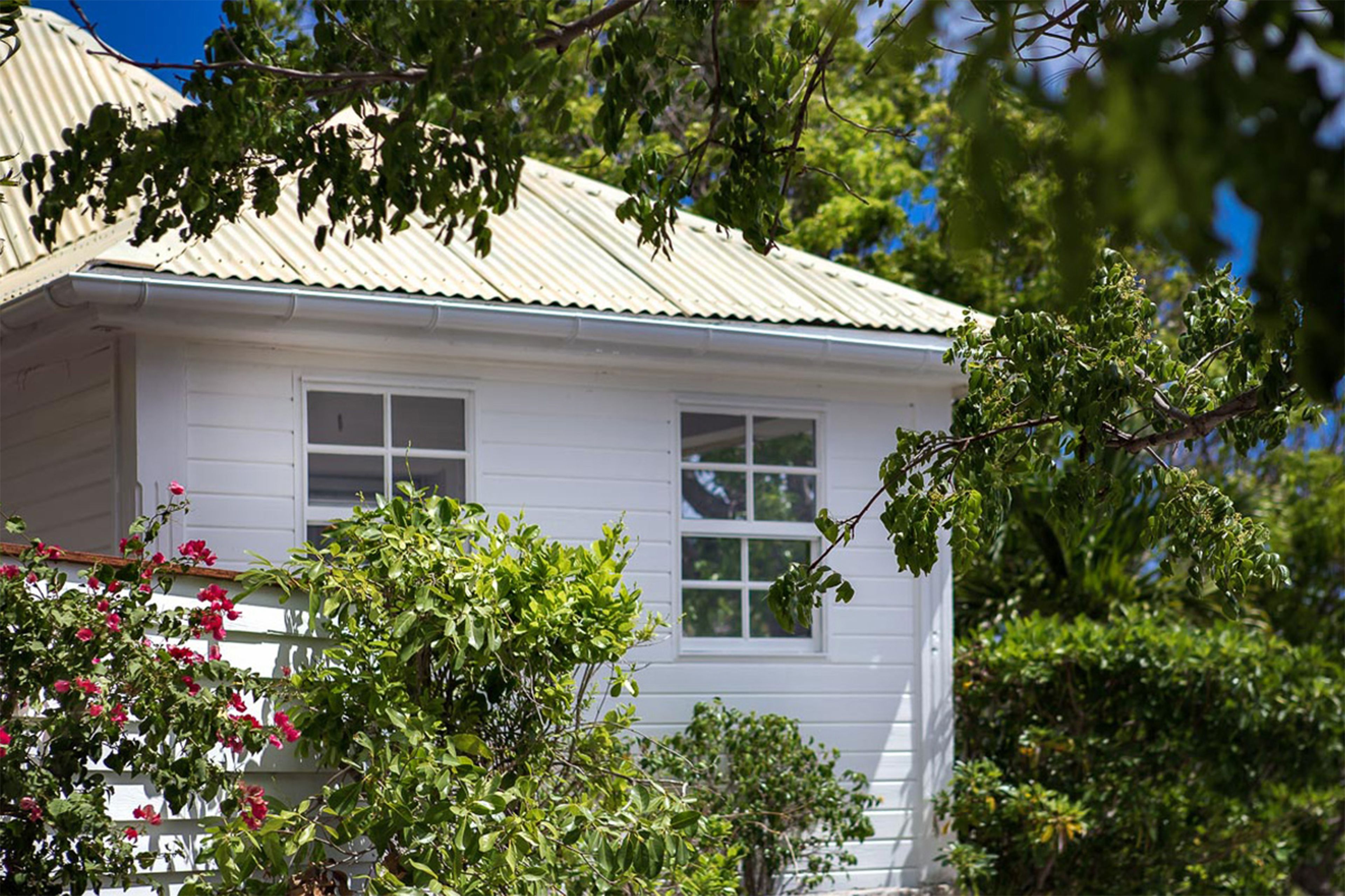 A white building with a corrugated metal roof, surrounded by lush greenery and flowering plants.