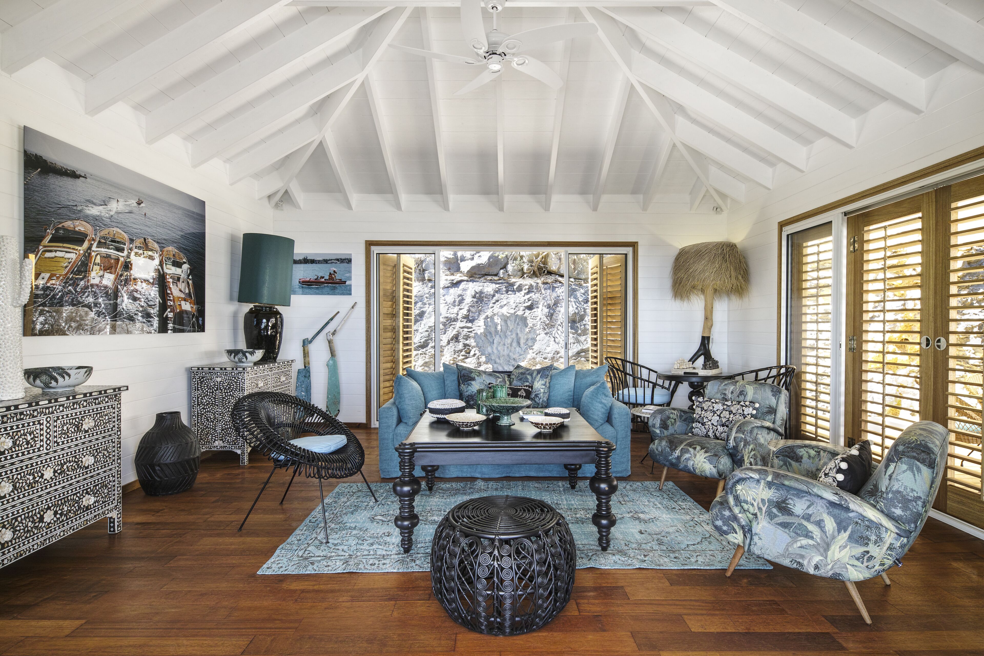 A spacious living room with a high ceiling, wooden flooring, a large window with shutters, a blue sofa, a coffee table, and a decorative rug.