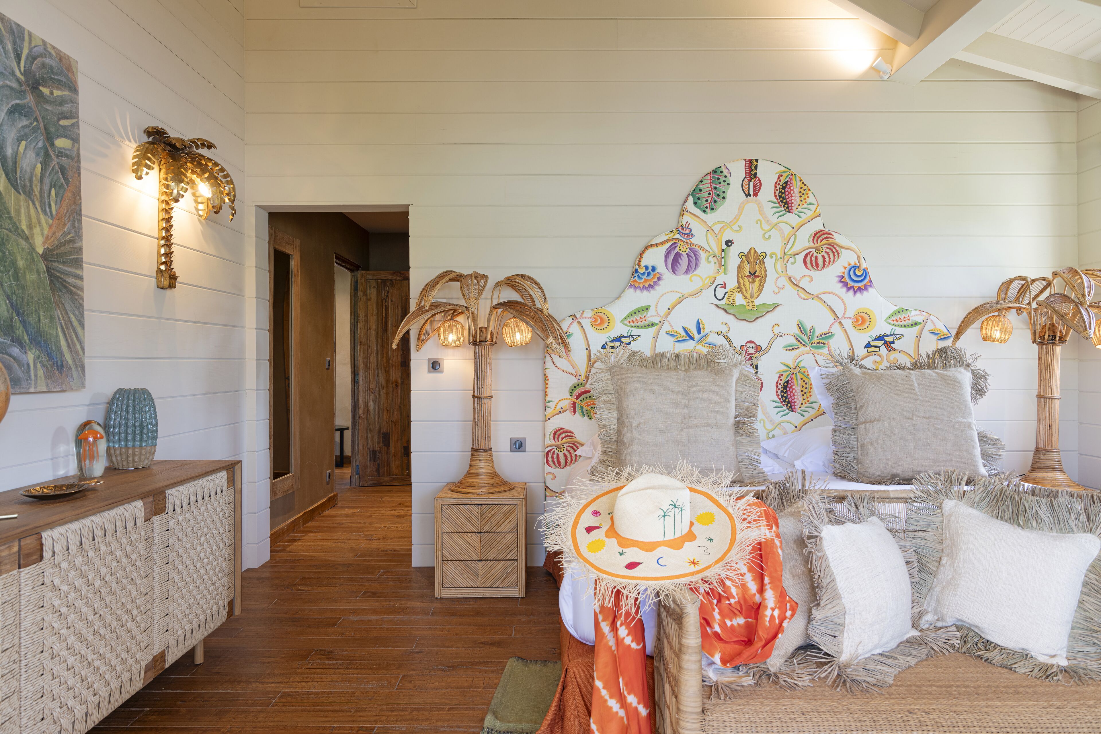 A cozy room with a wooden floor, a woven bench, and a decorative wall with a colorful mural. There are two wall-mounted lamps and a small table with decorative items.