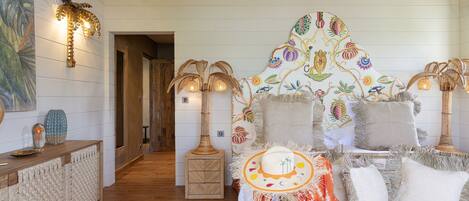 A cozy room with a wooden floor, a woven bench, and a decorative wall with a colorful mural. There are two wall-mounted lamps and a small table with decorative items.