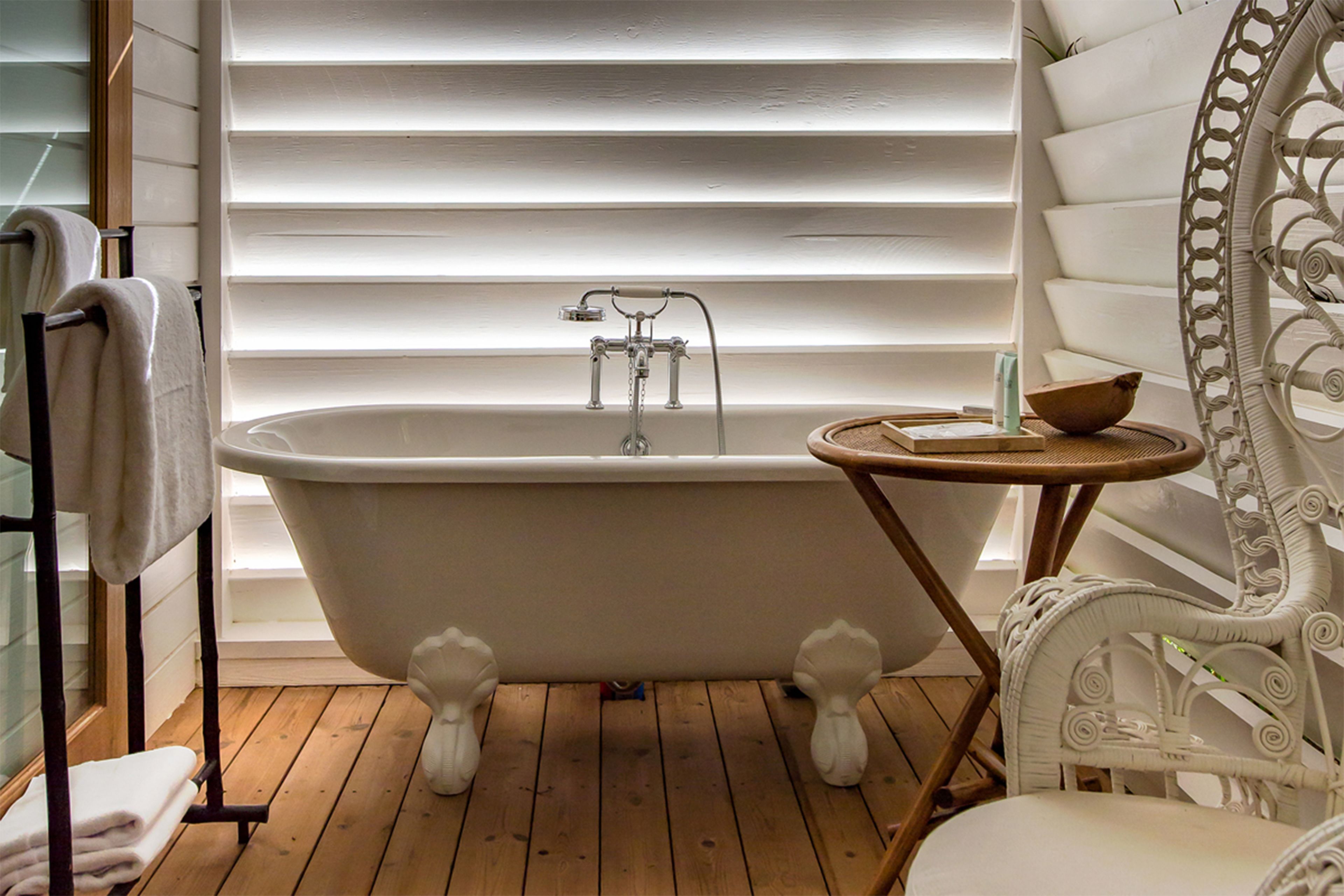 Une salle de bain avec une baignoire sur pieds, un plancher en bois, une petite table ronde avec un récipient, et une chaise blanche.