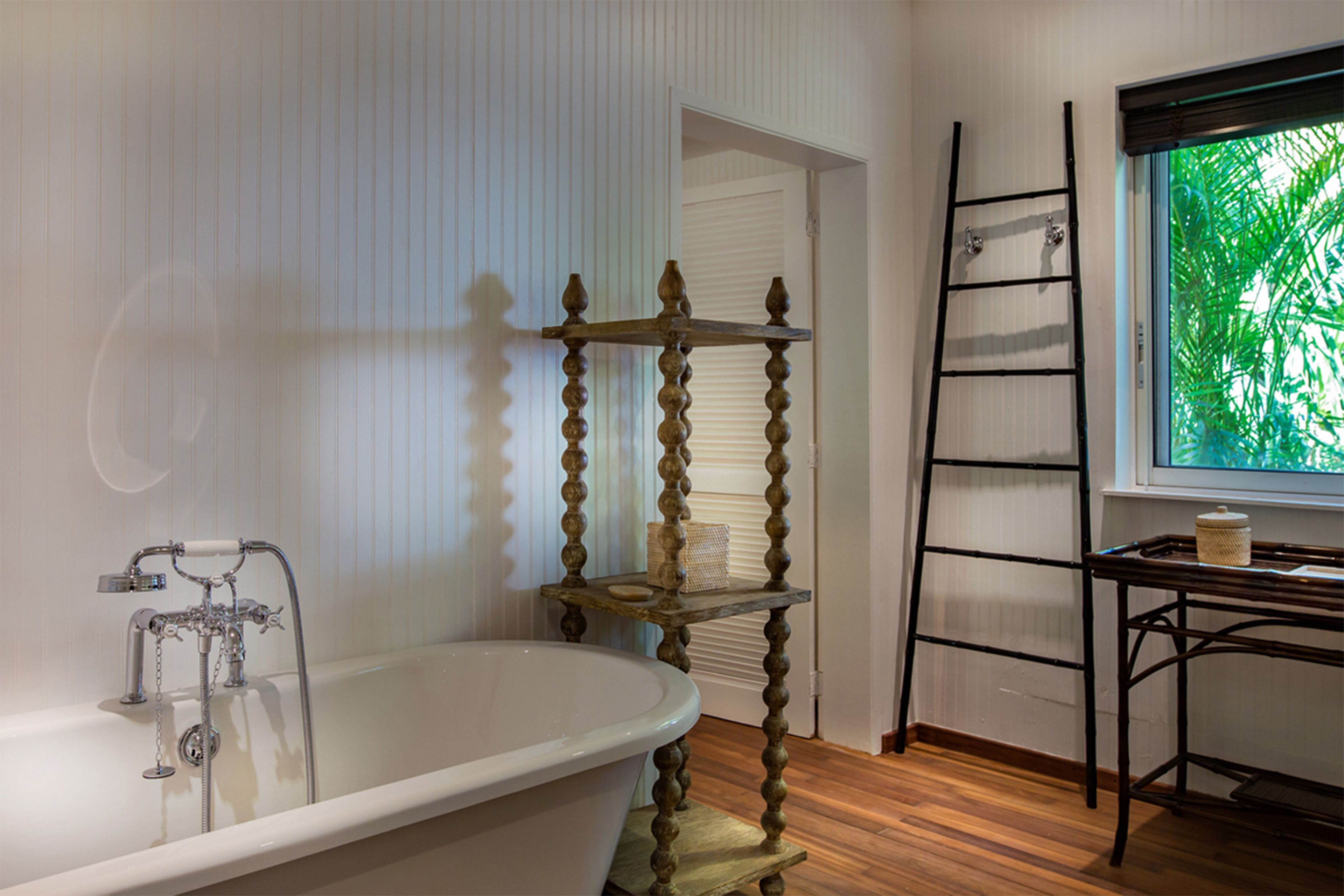 A bathroom with a freestanding bathtub, a wooden ladder, and a window with a view of green foliage.