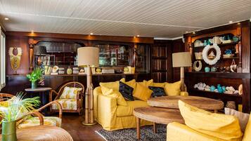 Uma sala de estar aconchegante com um sofá amarelo, cadeiras de vime, uma mesa redonda e uma estante cheia de objetos decorativos.