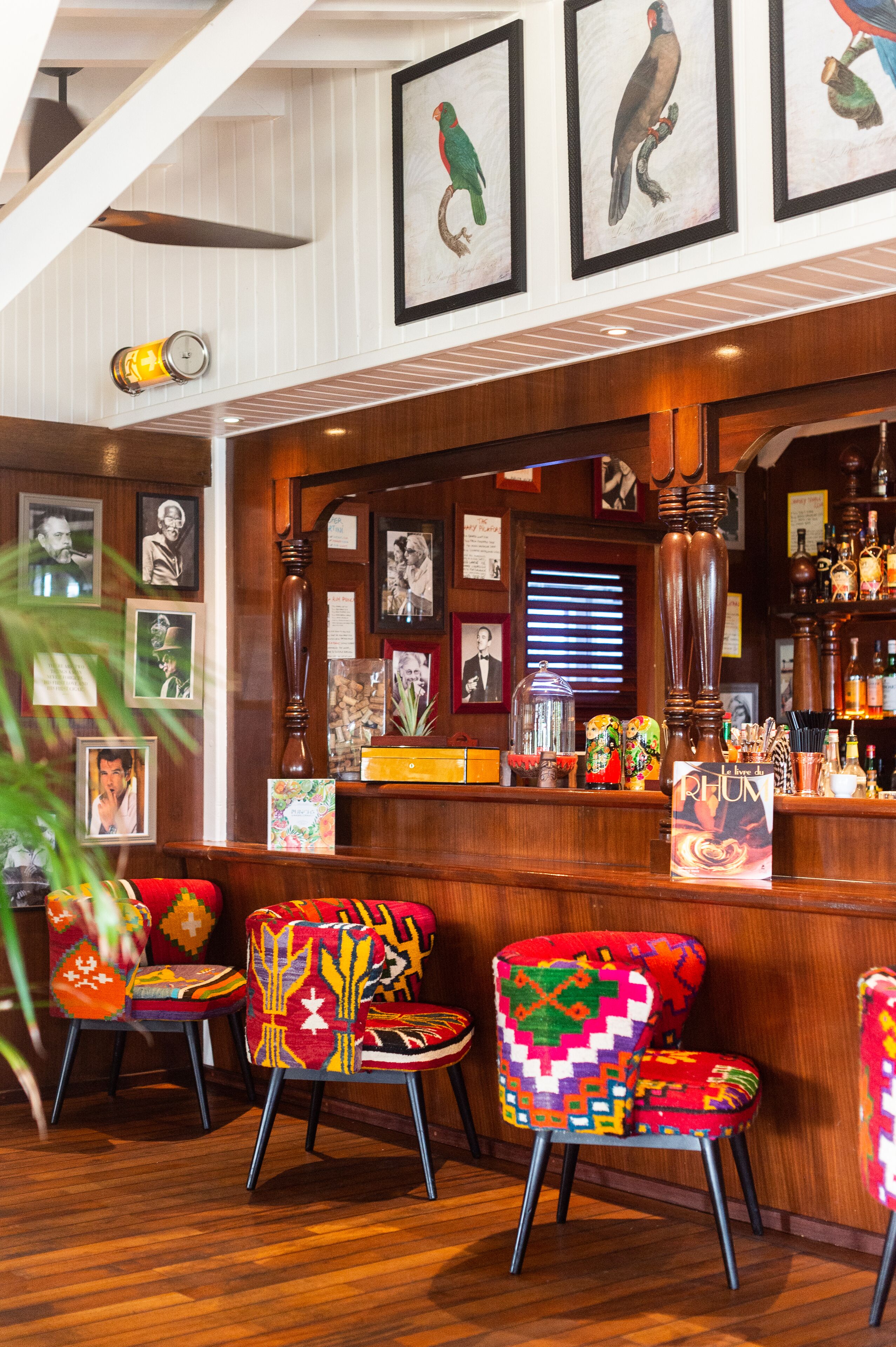 Una zona de bar con mobiliario de madera, sillas coloridas, cuadros enmarcados en la pared y una variedad de botellas en estanterías.