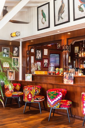 A bar area with wooden furniture, colorful chairs, framed pictures on the wall, and a variety of bottles on shelves.