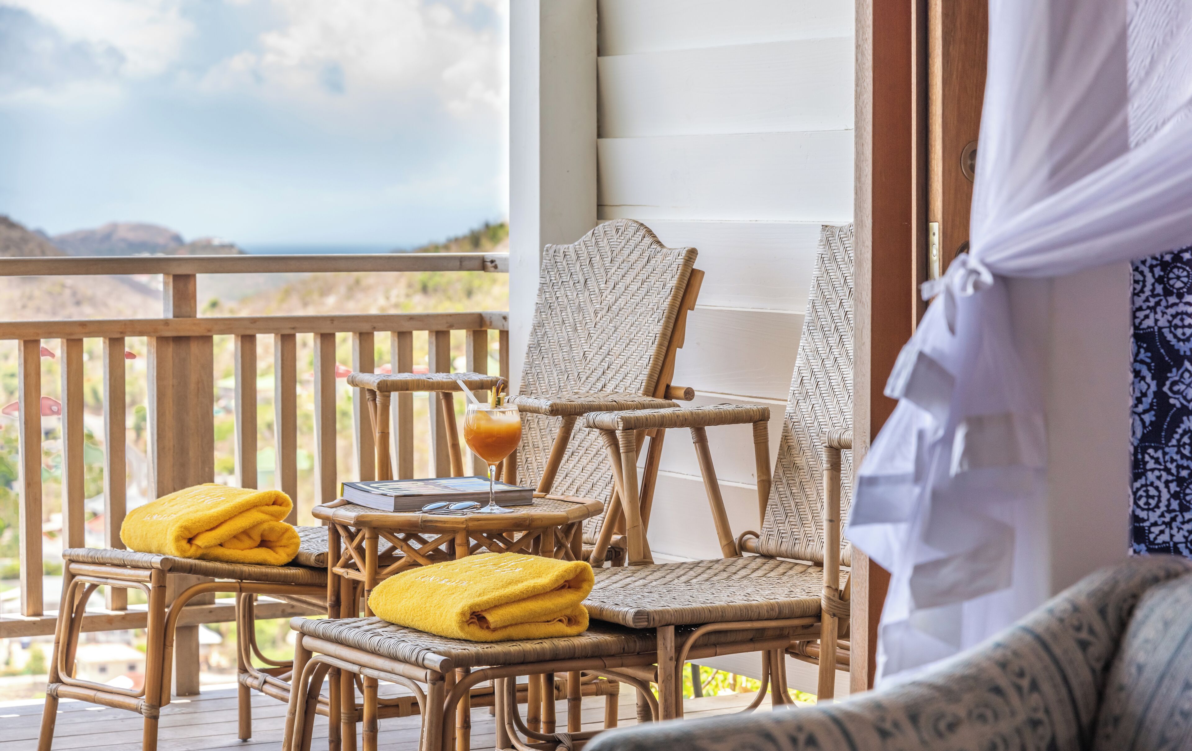 A wicker chair with a towel, a glass of orange juice, and a book on a wooden table on a balcony with a view of the ocean.