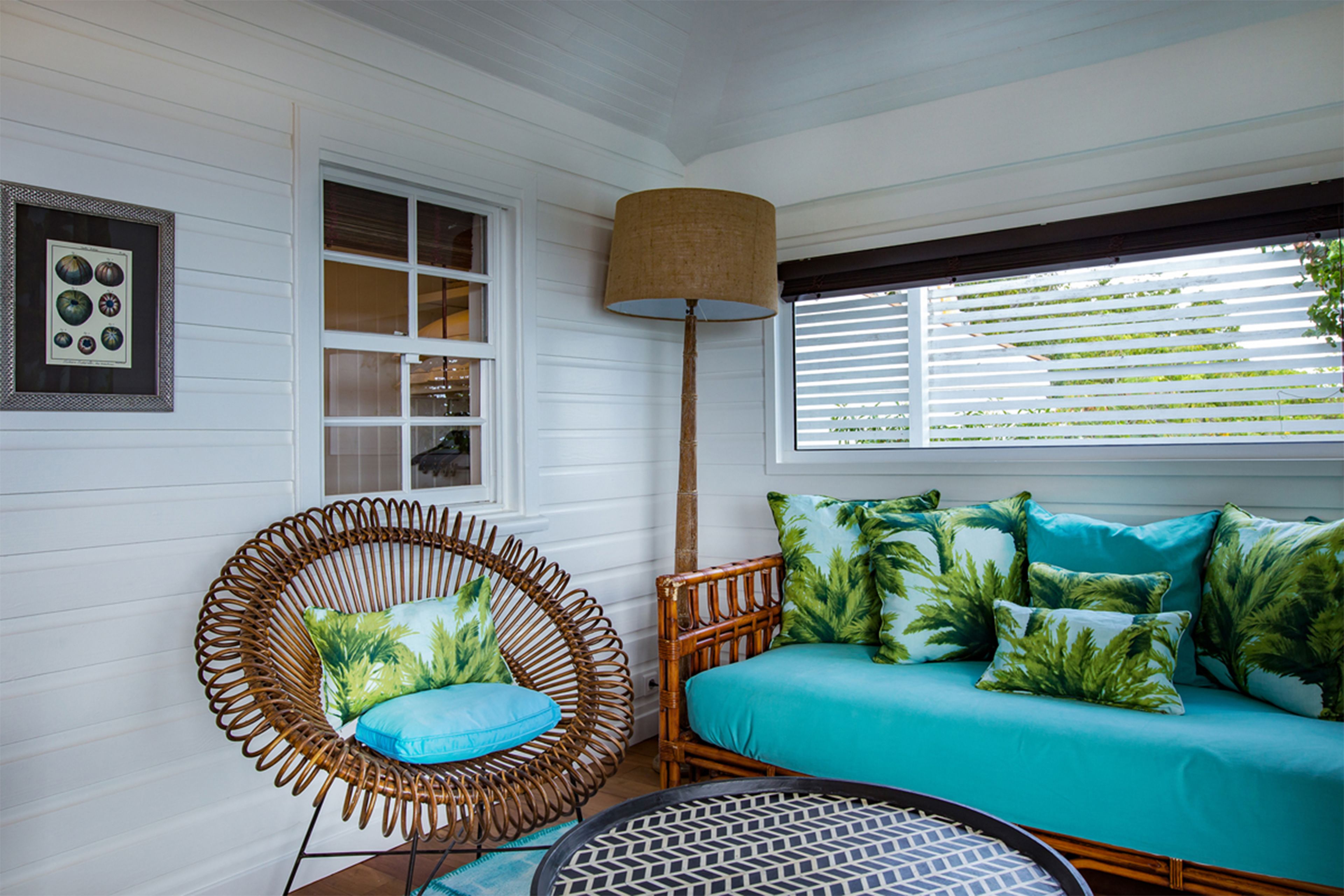 A room with a wicker chair, a sofa with tropical pillows, a floor lamp, and a window with blinds.