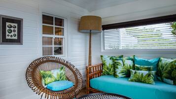 A room with a wicker chair, a sofa with tropical pillows, a floor lamp, and a window with blinds.