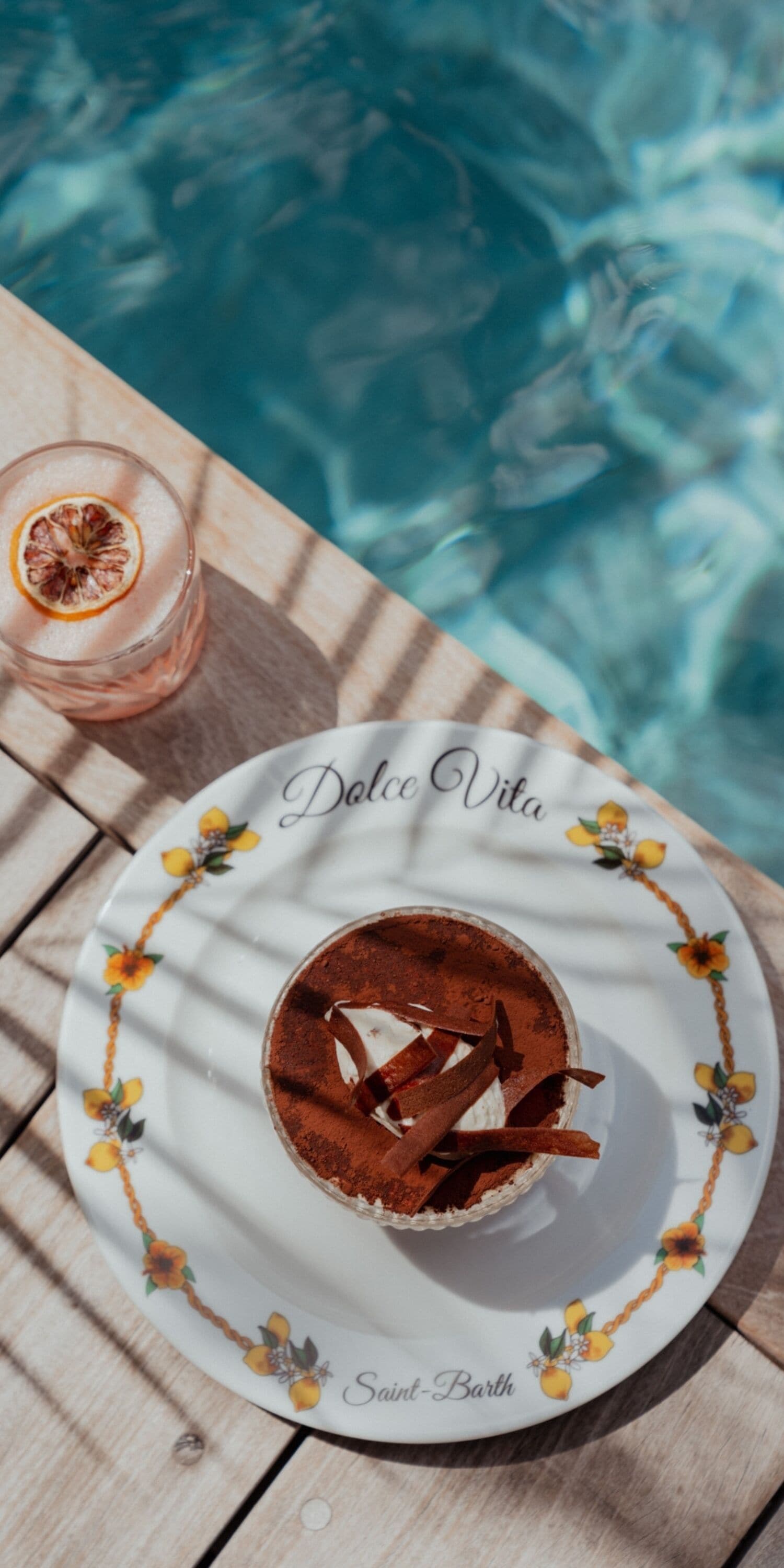 A dessert plate with a chocolate dessert and a glass of a pink drink, placed on a wooden table by a pool.