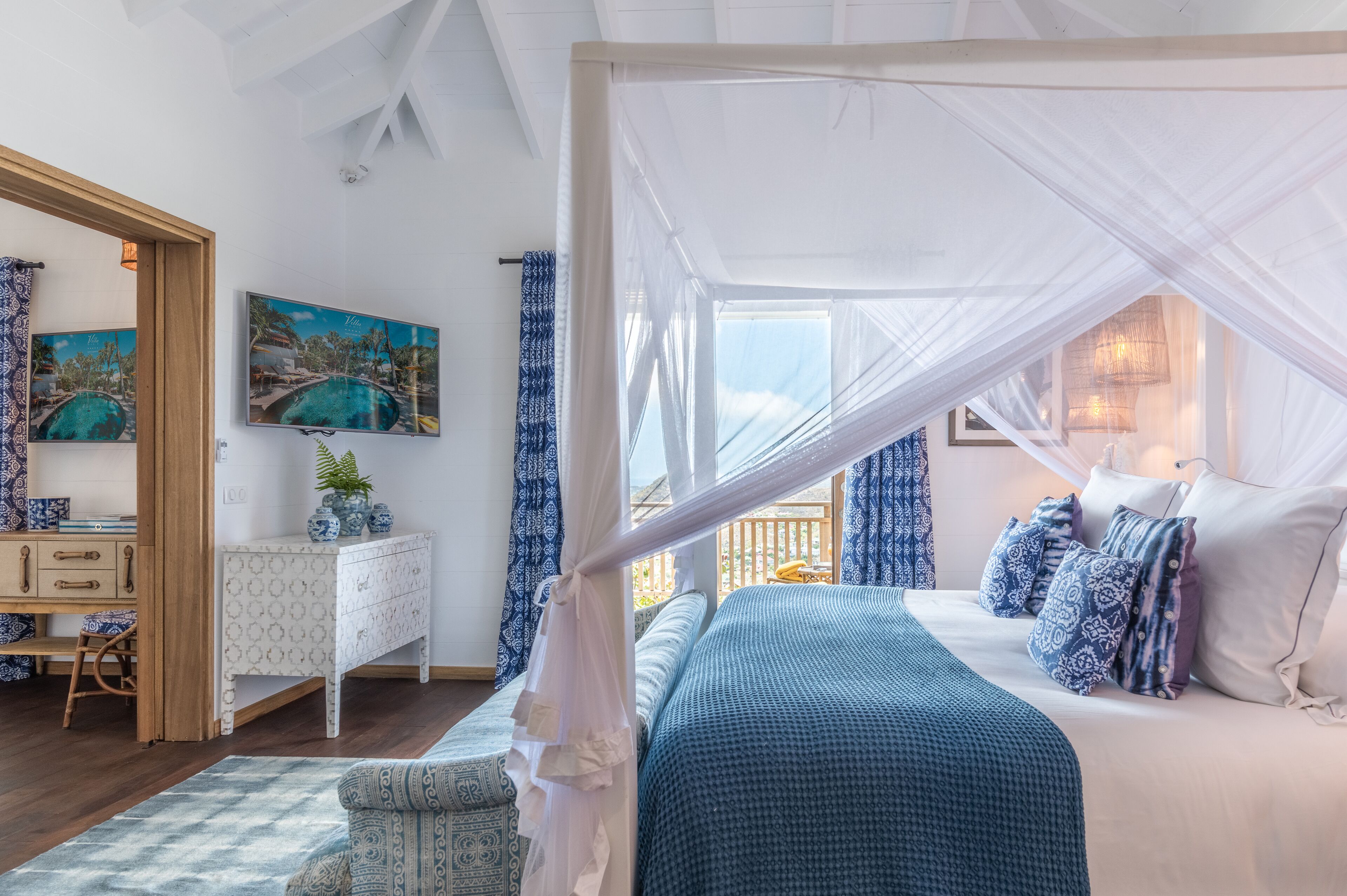 Ein Schlafzimmer mit Baldakine, einer weißen Kommode, einem kleinen Tisch mit Pflanze und einem Bild von einem Schwimmbad.